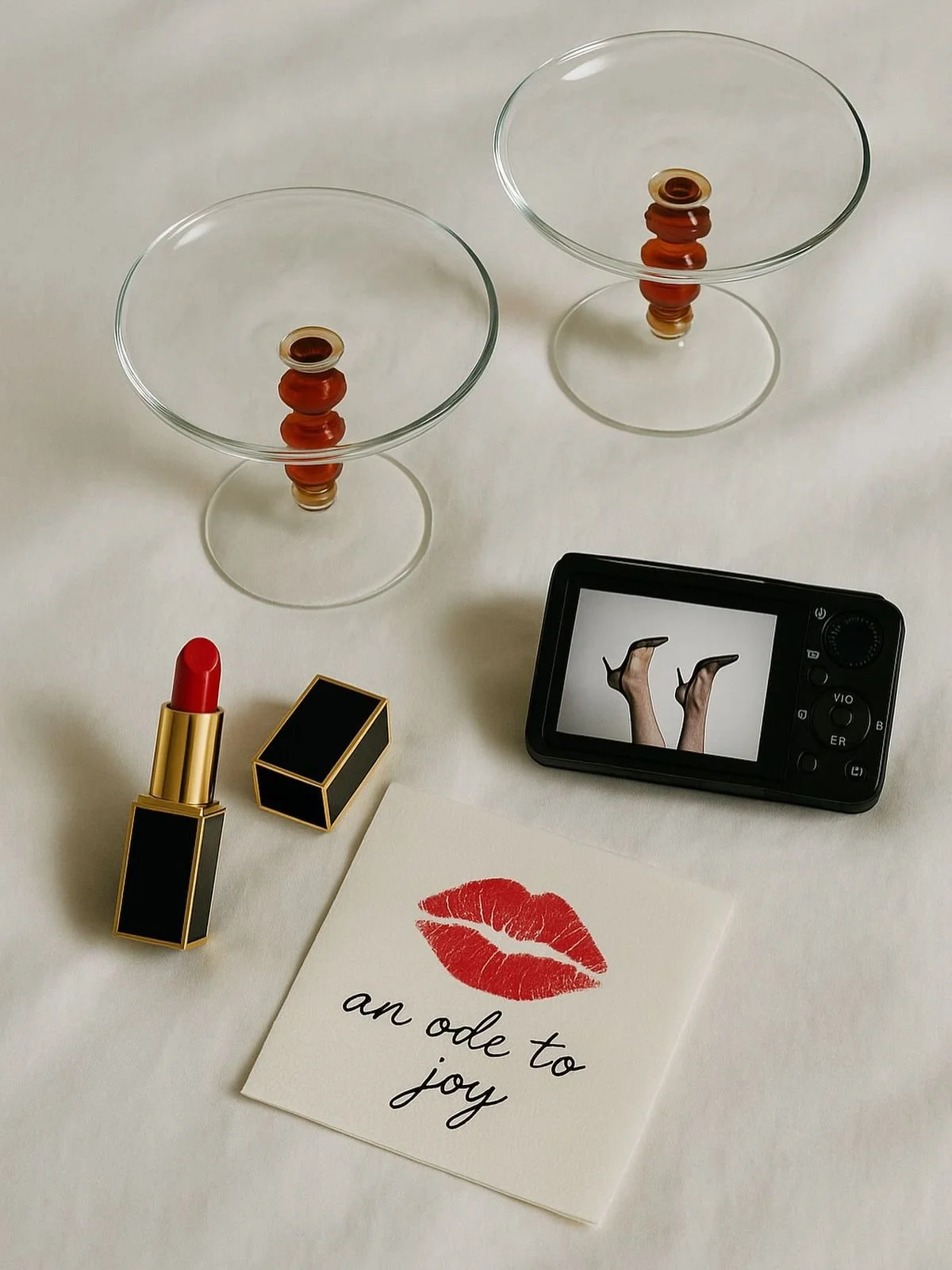 A flat lay of two empty glass dessert cups with orange and brown beads in the center, a black digital camera displaying legs in high heels, a red lipstick in a black and gold case, and a greeting card with red lipstick kiss mark and the phrase "an ode to joy" on a white surface.
