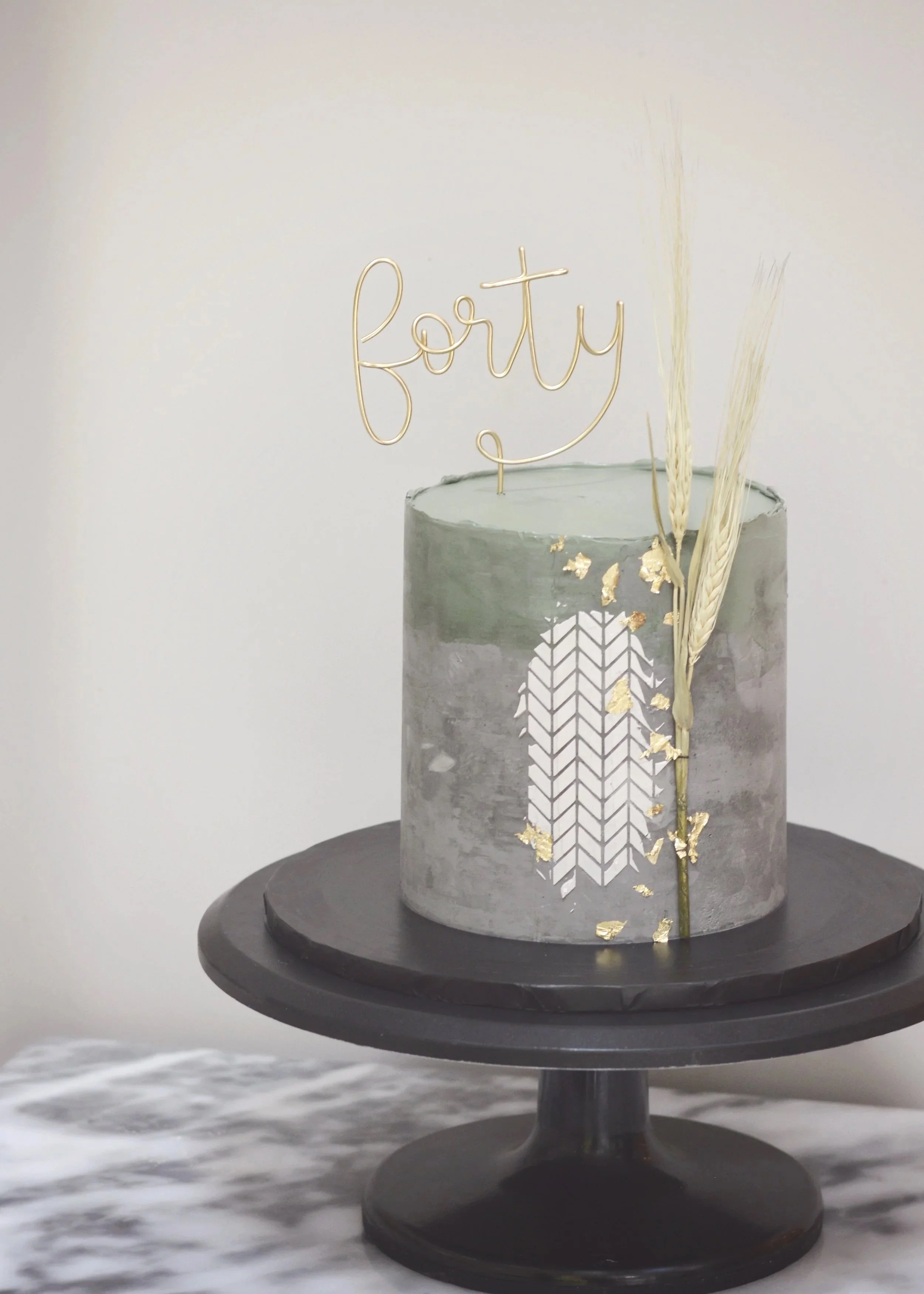  Grey Concrete effect celebration cake with olive colour buttercream decoration to the top half and a lighter colour cake stencil herringbone decoration and a barley branch decoration with a forty word cake topper in golden wire.  