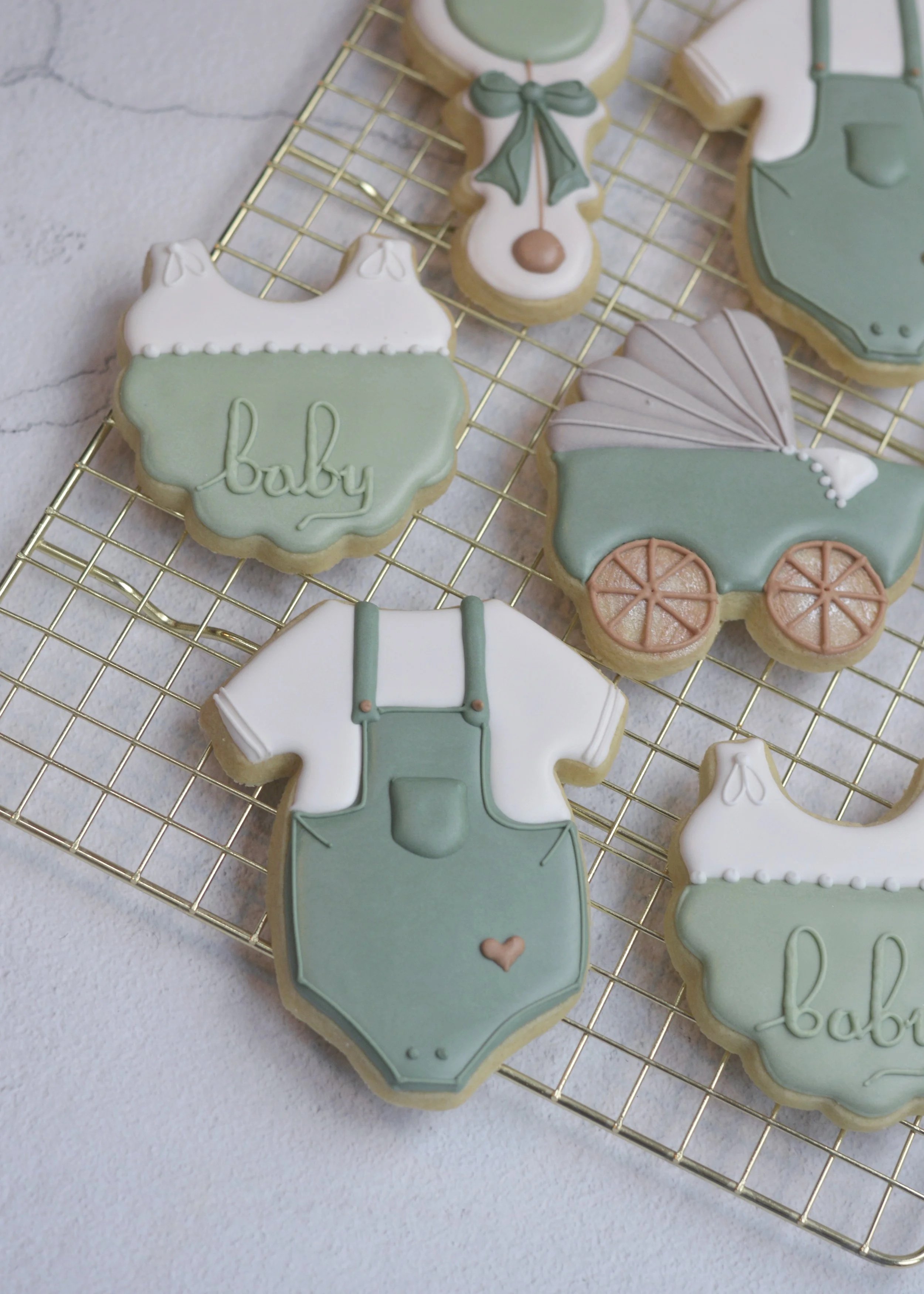 Gender neutral baby shower decorated cookies with pastel green, white, and beige colours, including a onesie, a vintage baby carriage, baby bottle, and the word 'baby' written on a bib cookie, displayed on a wire cooling rack.