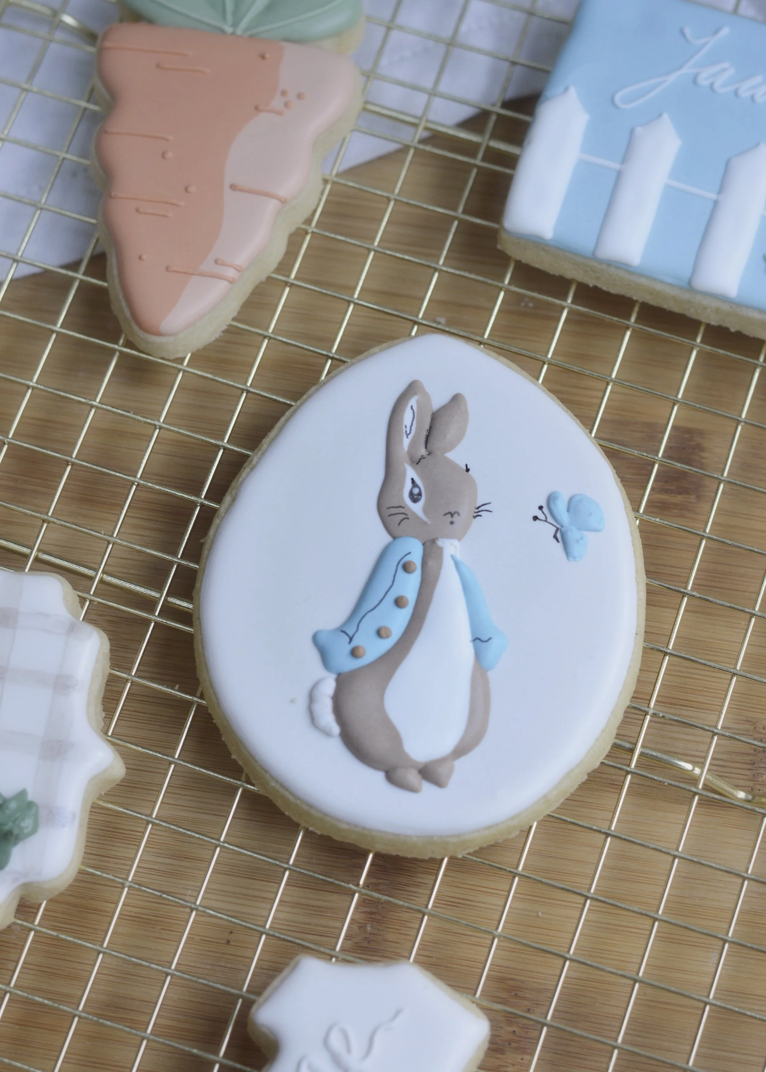 Decorated cookies on a wire rack, including one with Peter Rabbit wearing a blue jacket and a blue butterfly, and others with orange, blue, and white icing designs.
