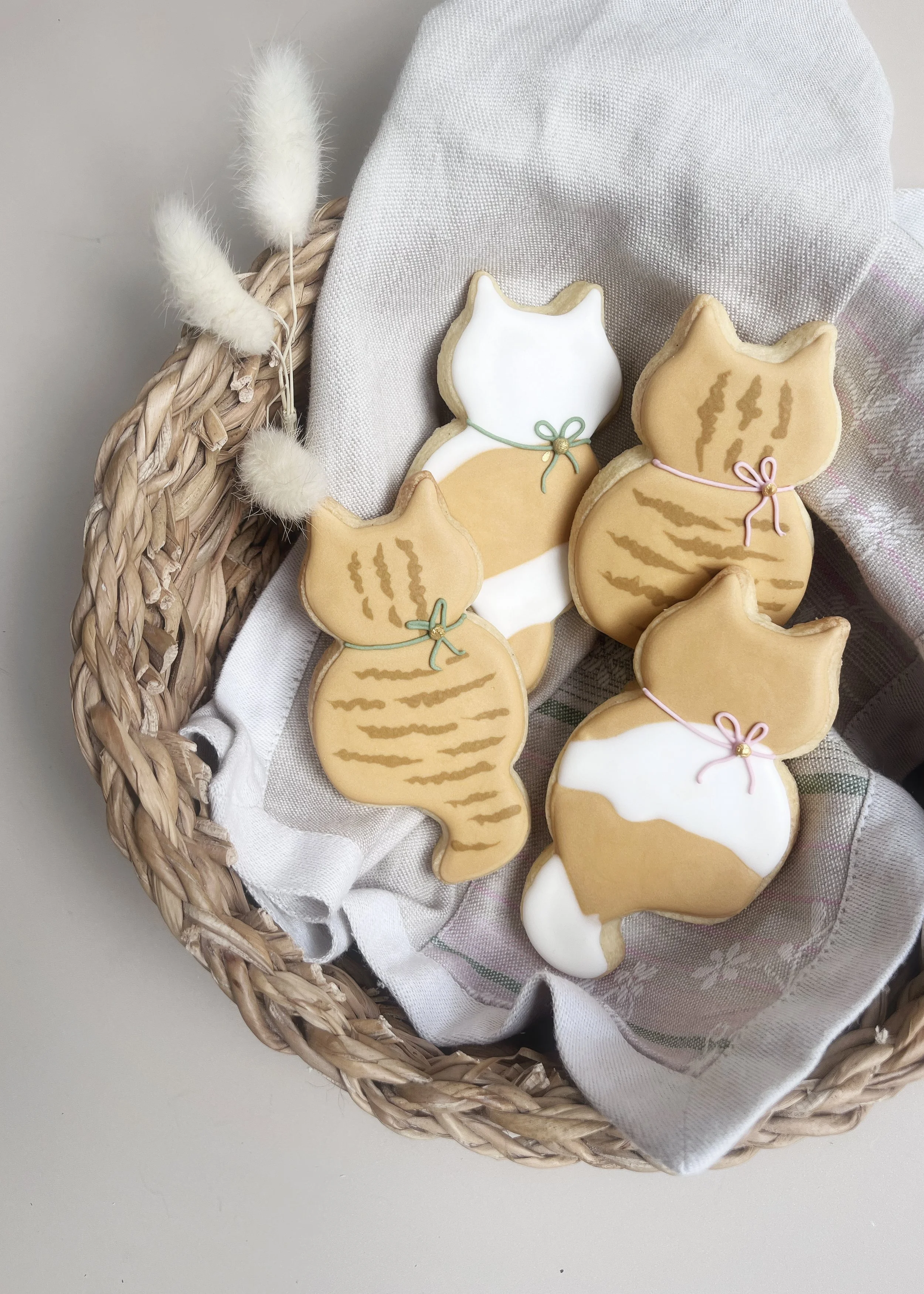 Cookies decorated to look like cats in a basket with a cloth, some with white icing and others with tan icing, each with a small ribbon around their necks.