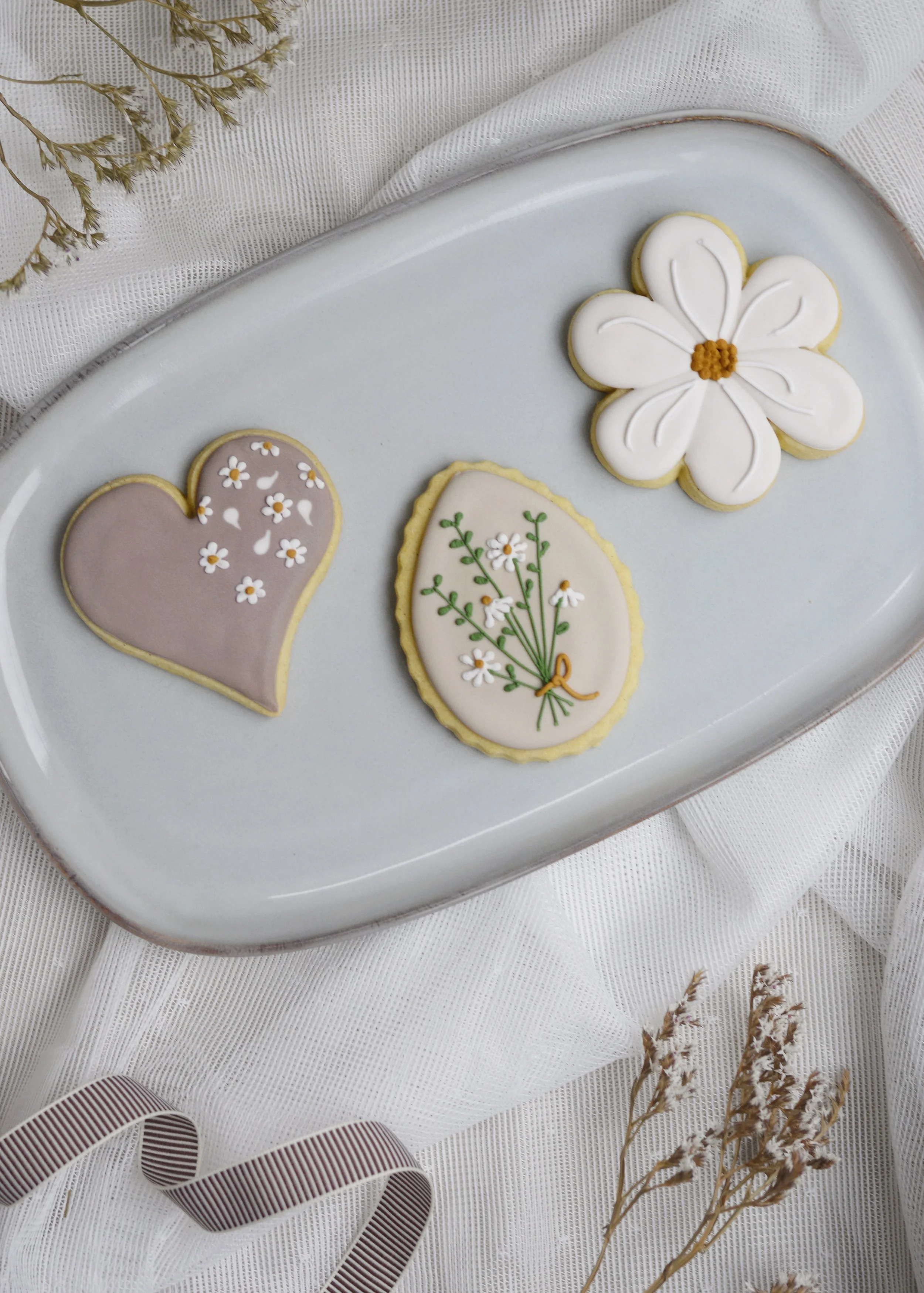 Decorative cookies on a white oval plate, including a heart-shaped cookie with white daisies, an oval cookie with daisies and green leaves tied with an orange ribbon, and a flower-shaped cookie with white icing and an orange center, surrounded by dried flowers and a white textured cloth.