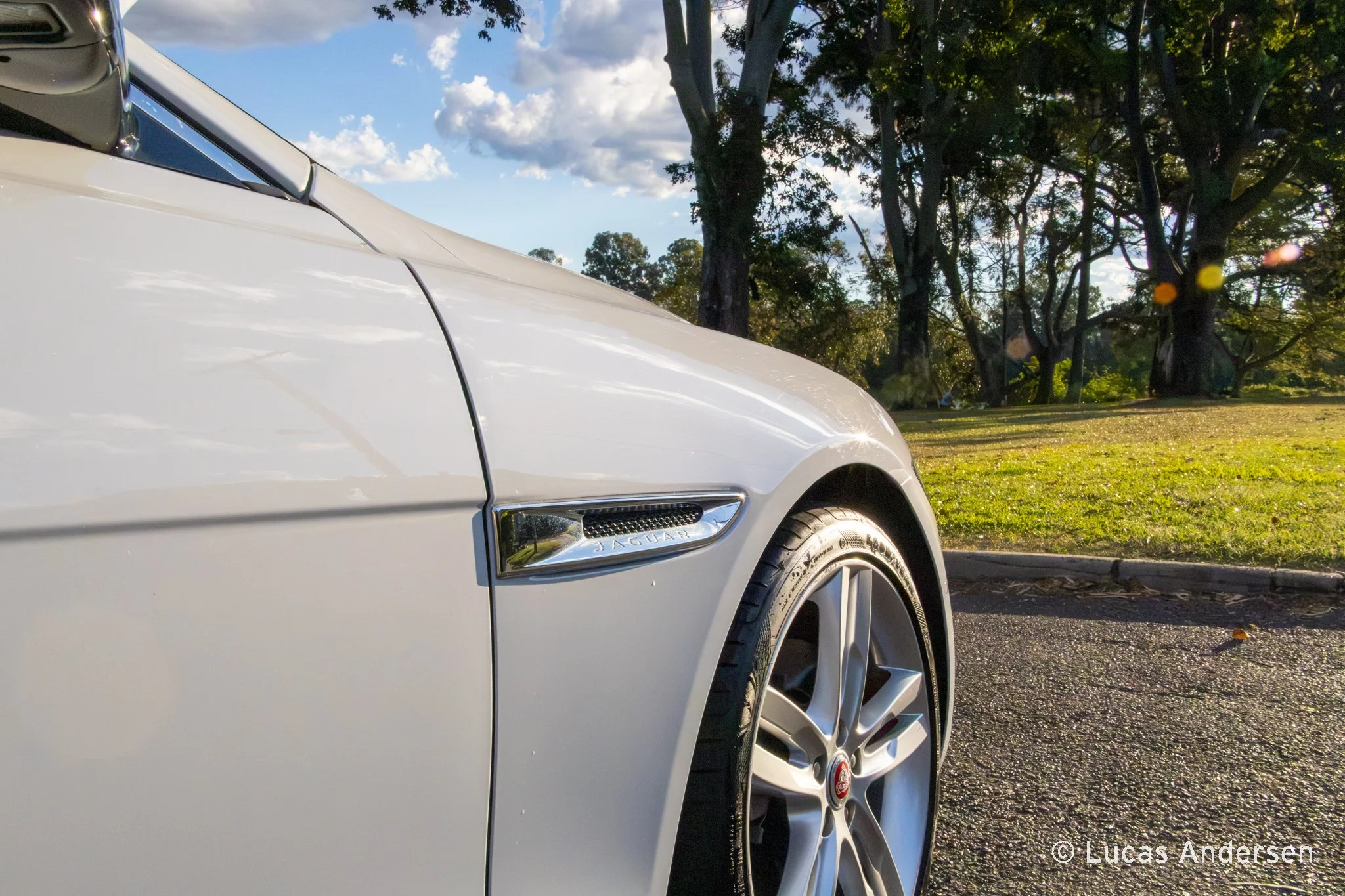 Close-up of the front left side of a white Jaguar car parked outdoors near a grassy area with trees, under partly cloudy sky.