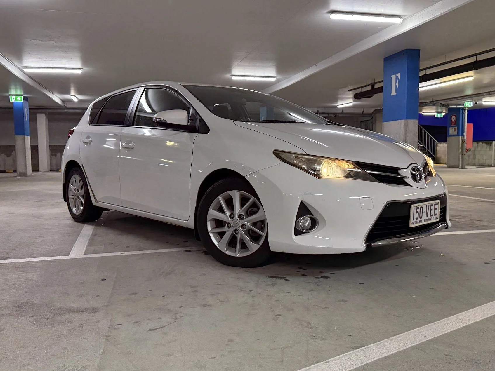 White Toyota hatchback car parked in a multi-level parking garage with bright overhead lights and blue structural columns.