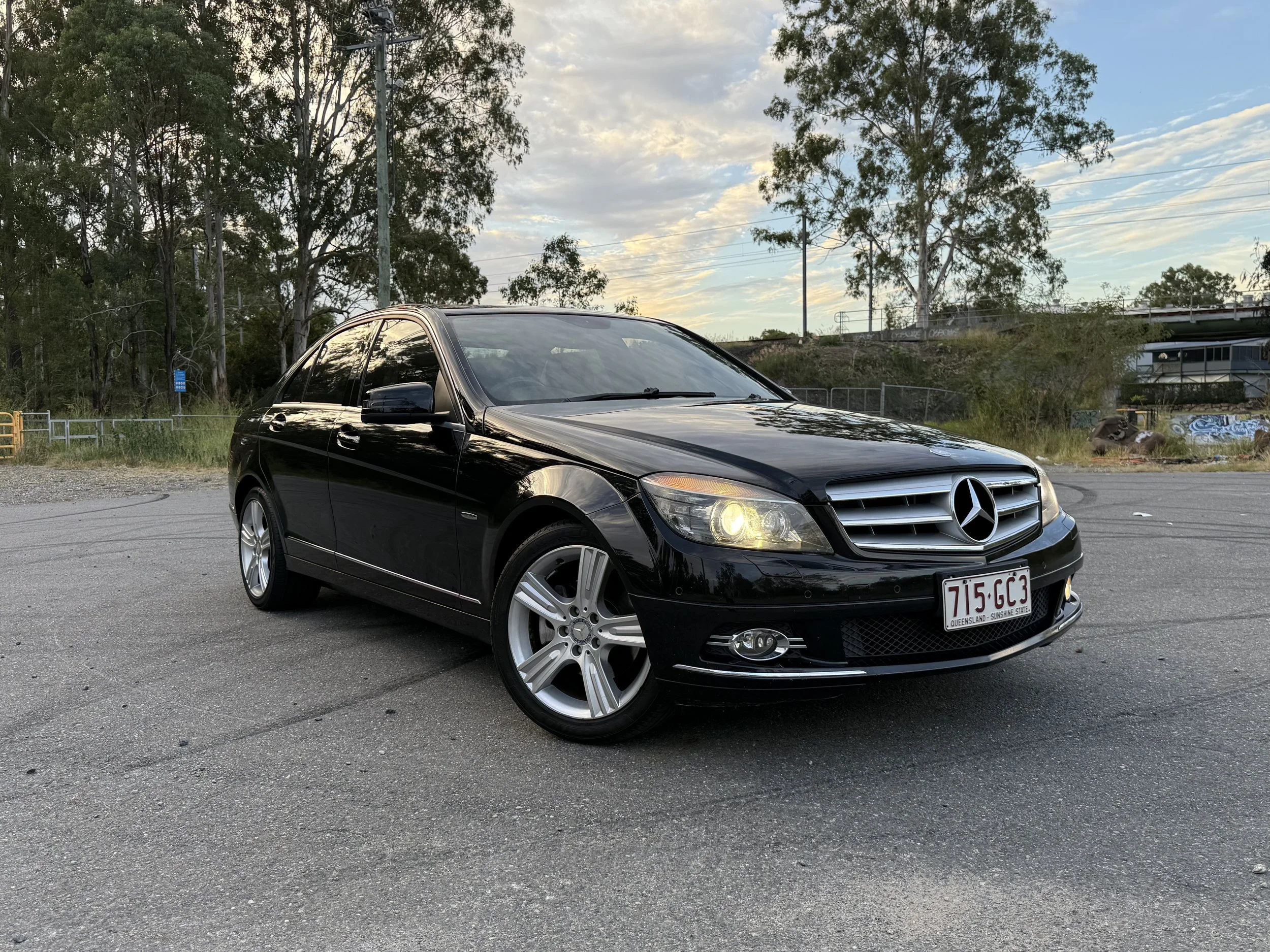 A black Mercedes-Benz car parked outdoors, with trees and utility poles in the background during sunset or sunrise.