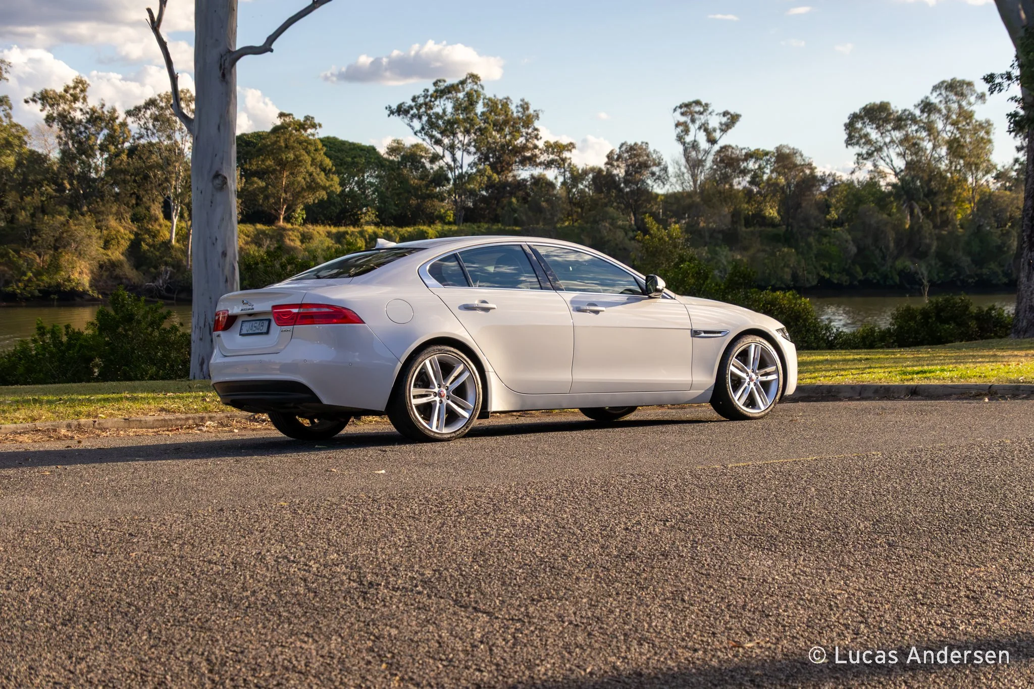A white luxury sedan car parked on the side of a road near a grassy area and trees with a river and blue sky with clouds in the background.