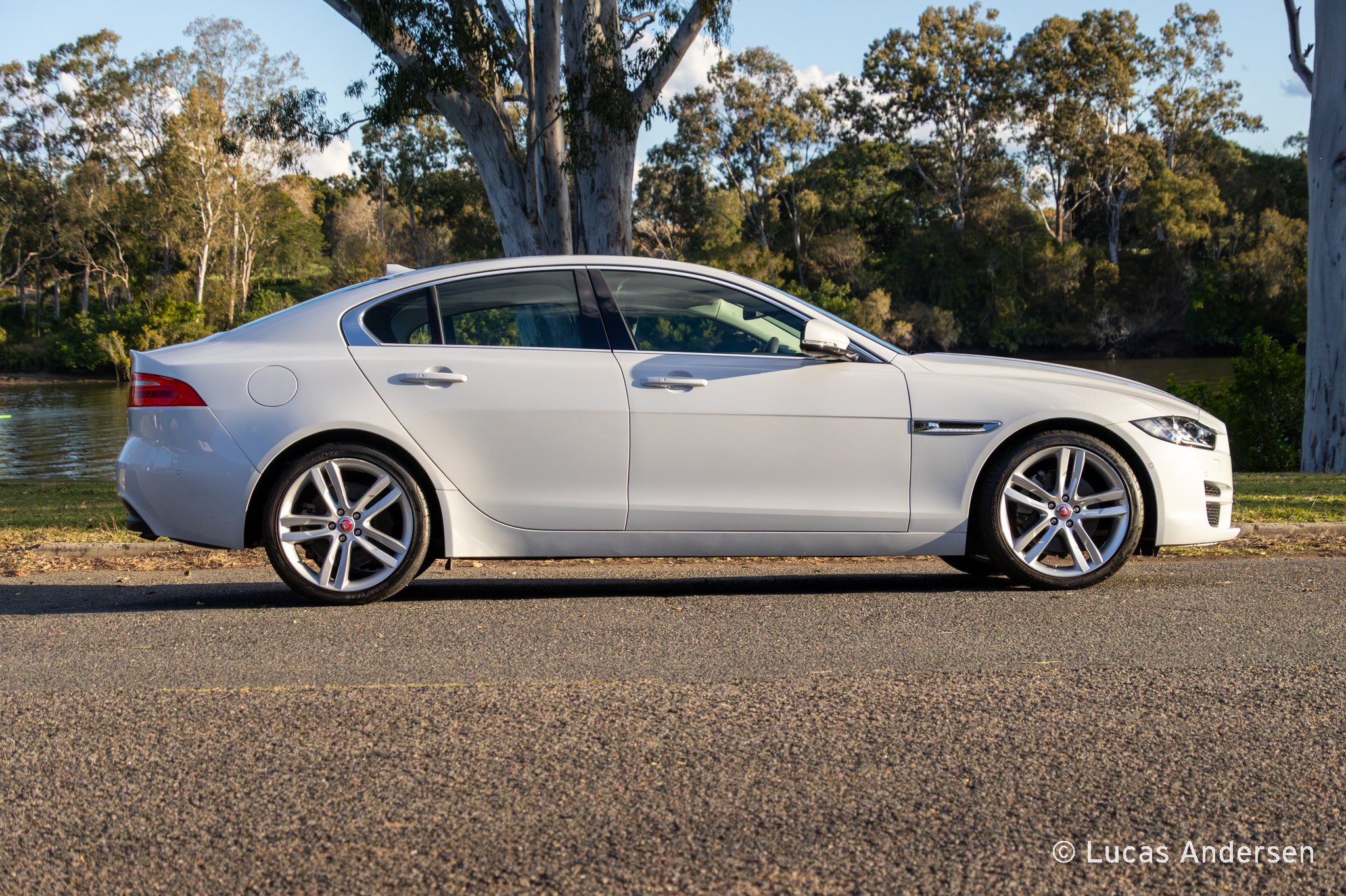 A white luxury sedan parked on a paved road with a backdrop of tall trees and a river.