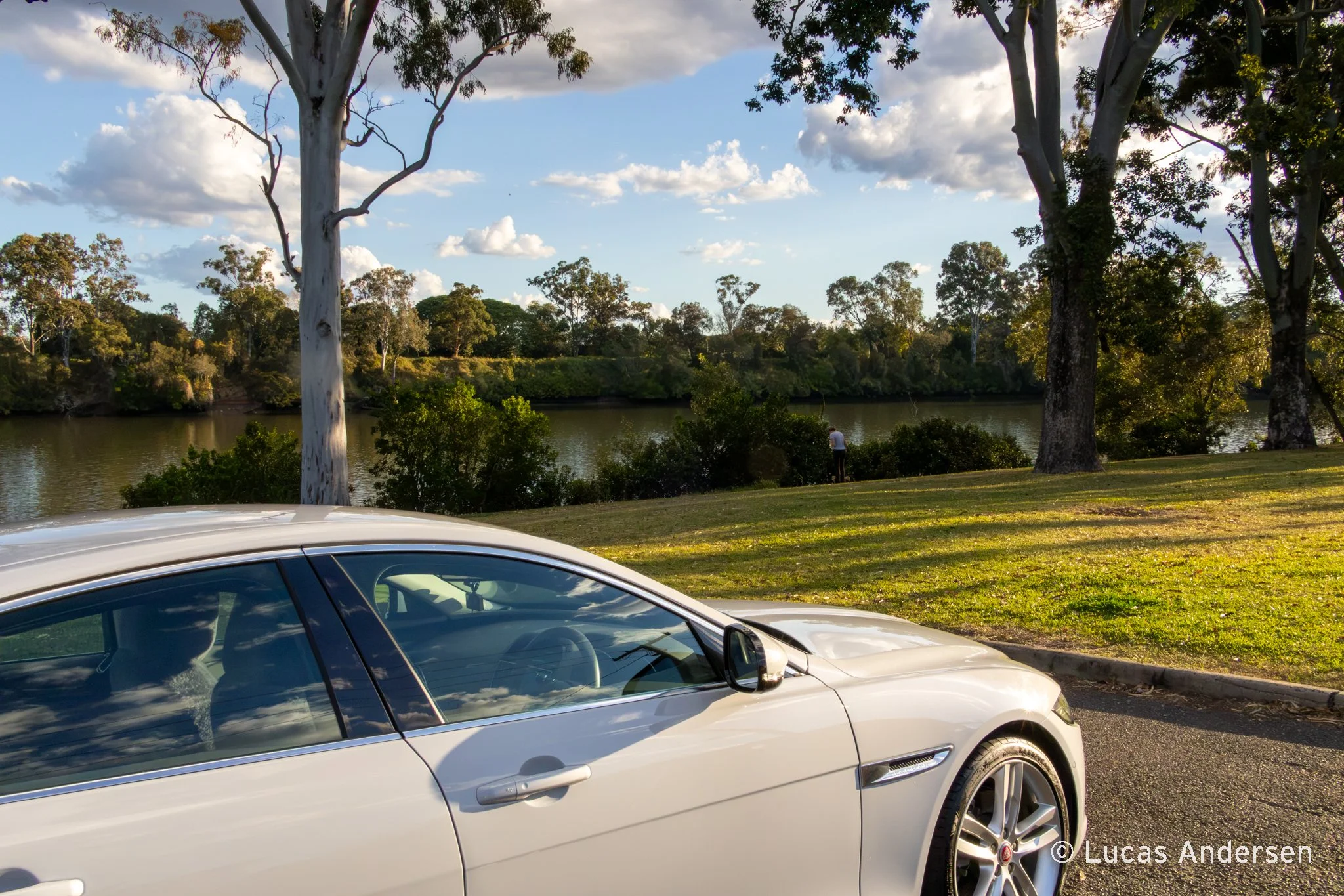 A white luxury car parked in front of a river and trees in a park during the daytime, with a partly cloudy sky.