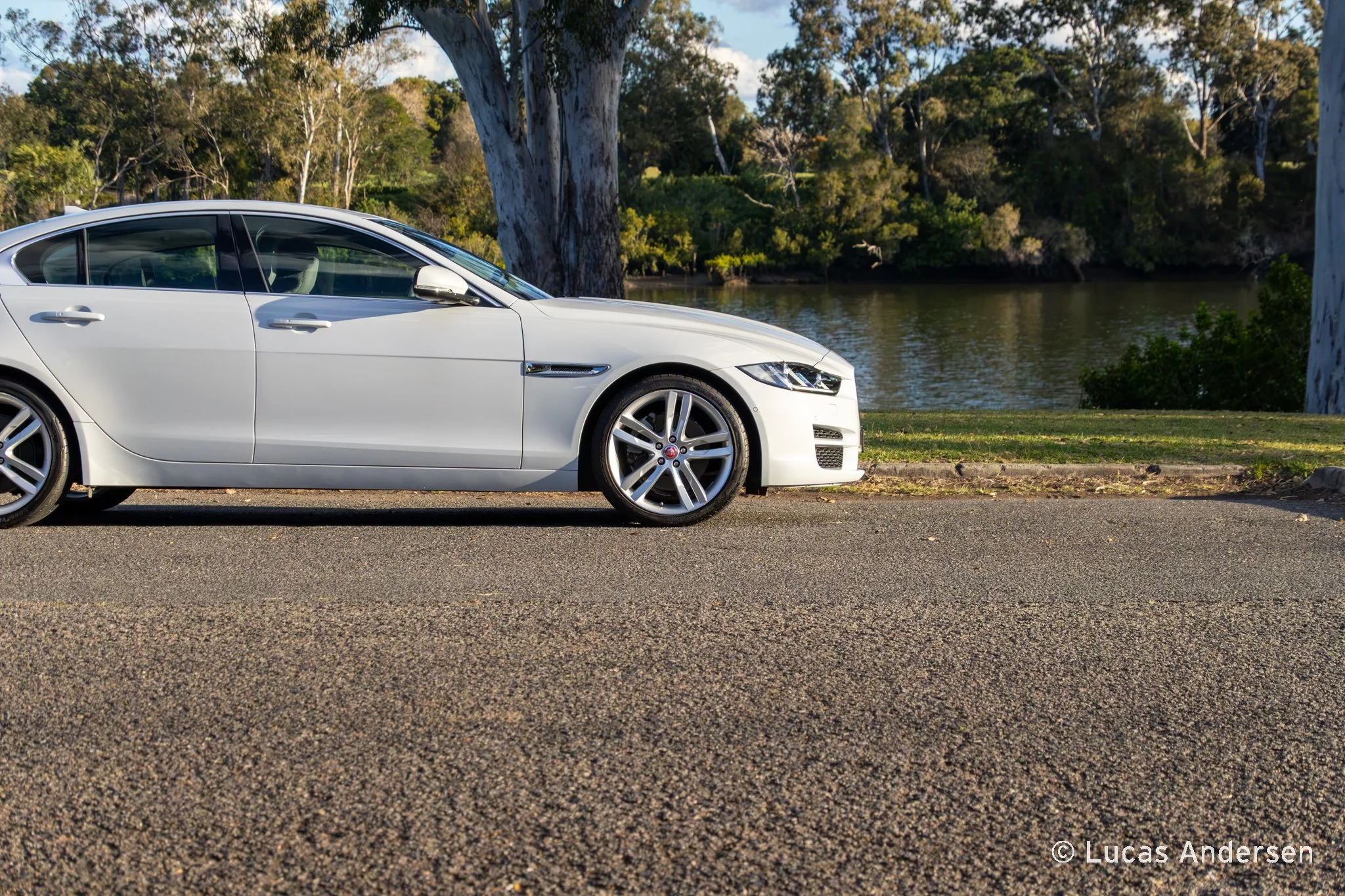 A white luxury sedan car parked on a roadside near a body of water with trees and bushes in the background.