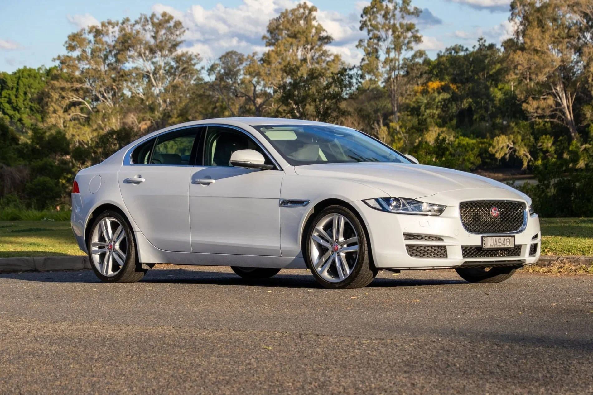 A white Jaguar sedan parked on a street with greenery and trees in the background.