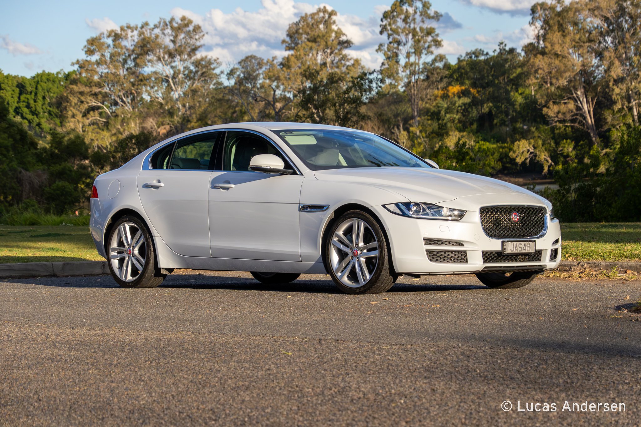A white Jaguar sedan parked on the side of a road with grass and trees in the background under a partly cloudy sky.