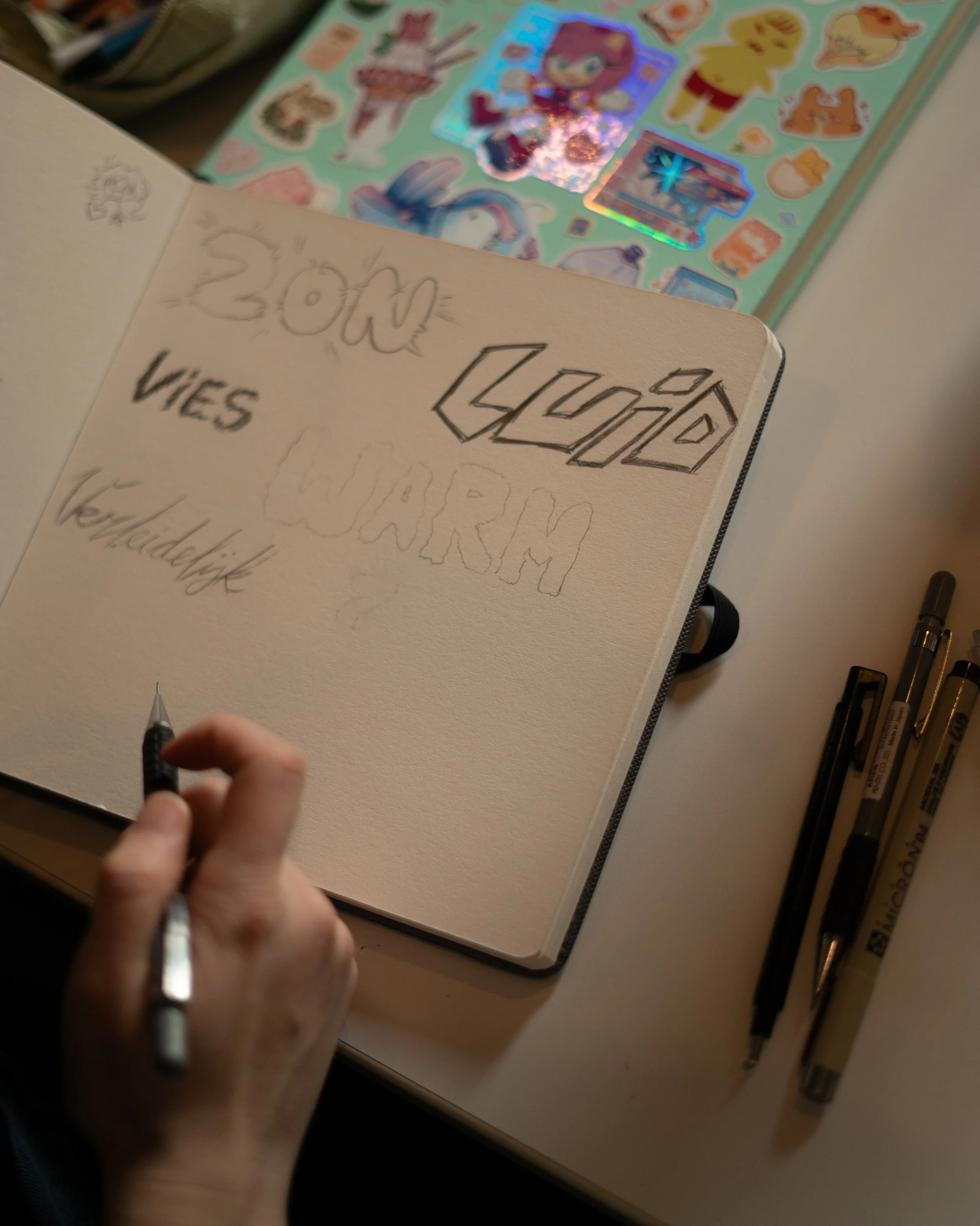 Een persoon tekent woorden en tekeningen in een schetsboek, met stickers en stiften op de tafel ernaast. Les striptekenen en animatie bij Skill School Ede.