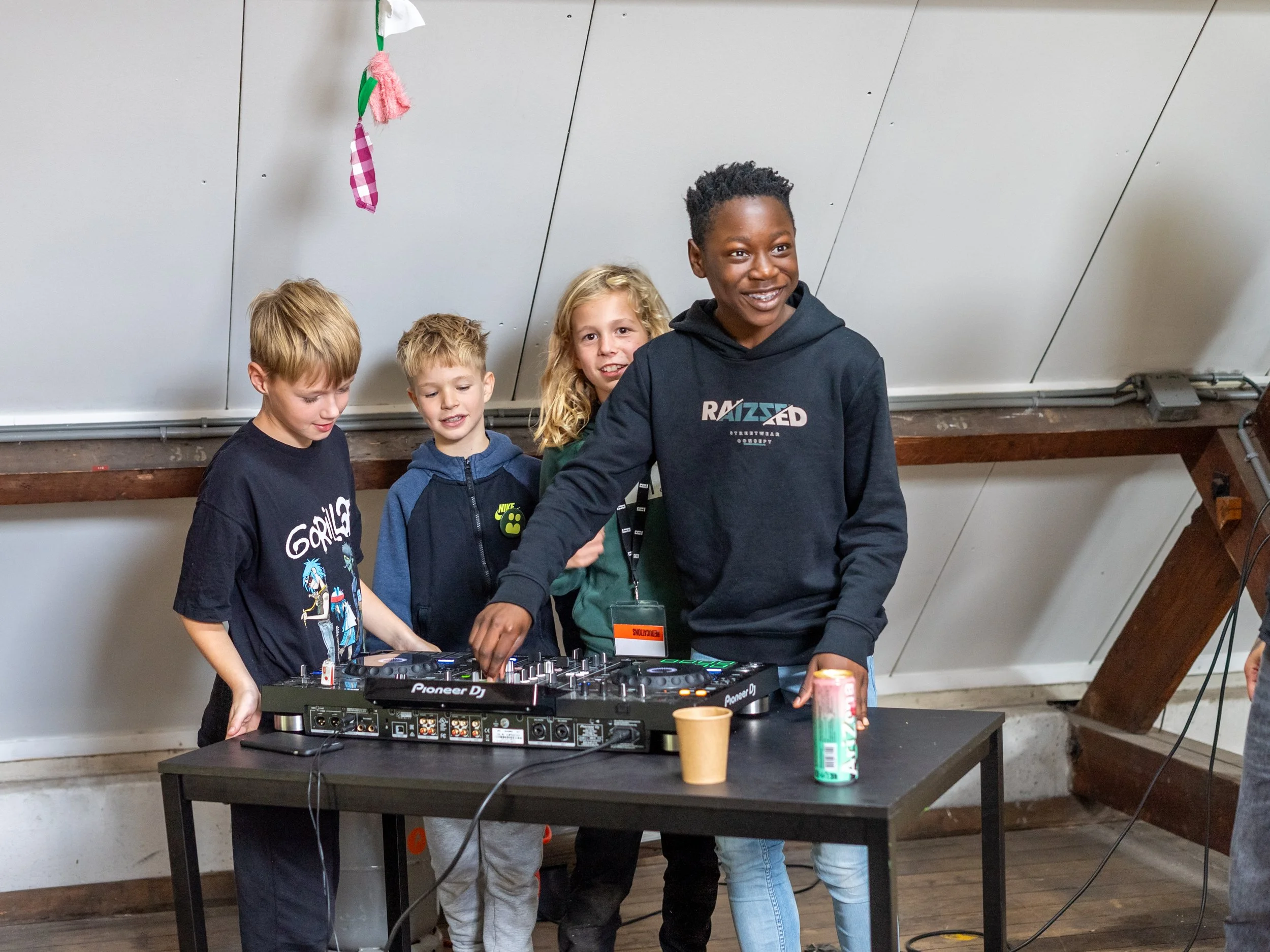 Vier kinderen staan achter een DJ-set, één kind bedient de apparatuur terwijl de andere drie toekijken. Ze zijn in een kamer met een schuine muur en houten structuur. Herfstweek bij Skill School junior in Ede