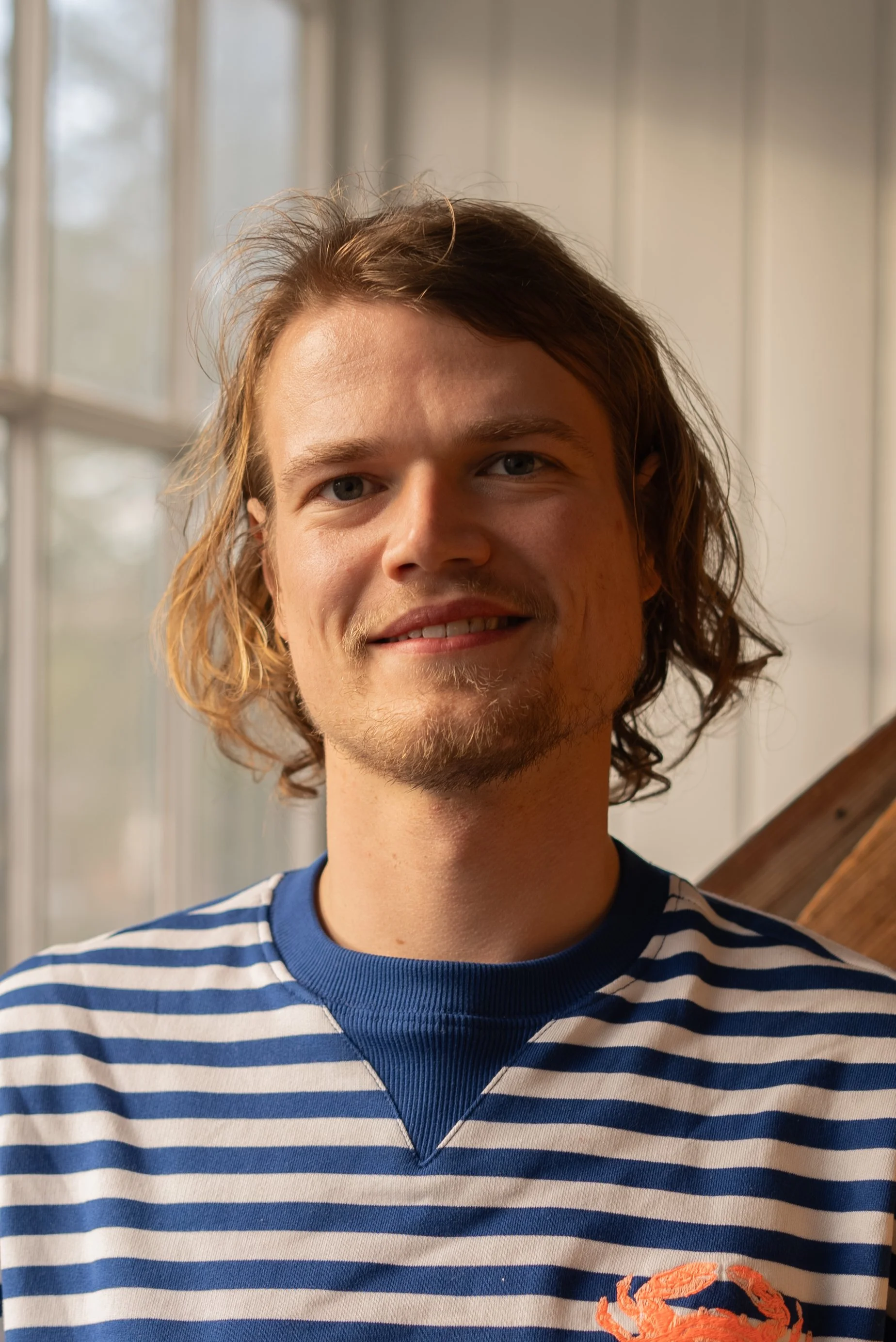 Portret van een jongeman met halflang golvend haar die binnen in een kamer staat, gekleed in een blauwe en witte gestreepte trui, met grote ramen en houten trap op de achtergrond. Docent bij Skill School Ede.