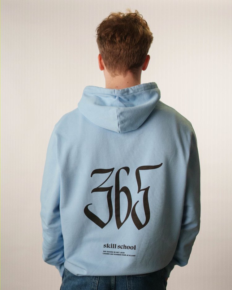 Persoon met korte, golvende bruin haar die een lichtblauwe hoodie draagt met een grote '365' op de rug en de woorden 'Skill School' onderin, staand tegen een witte achtergrond. Skill School merch