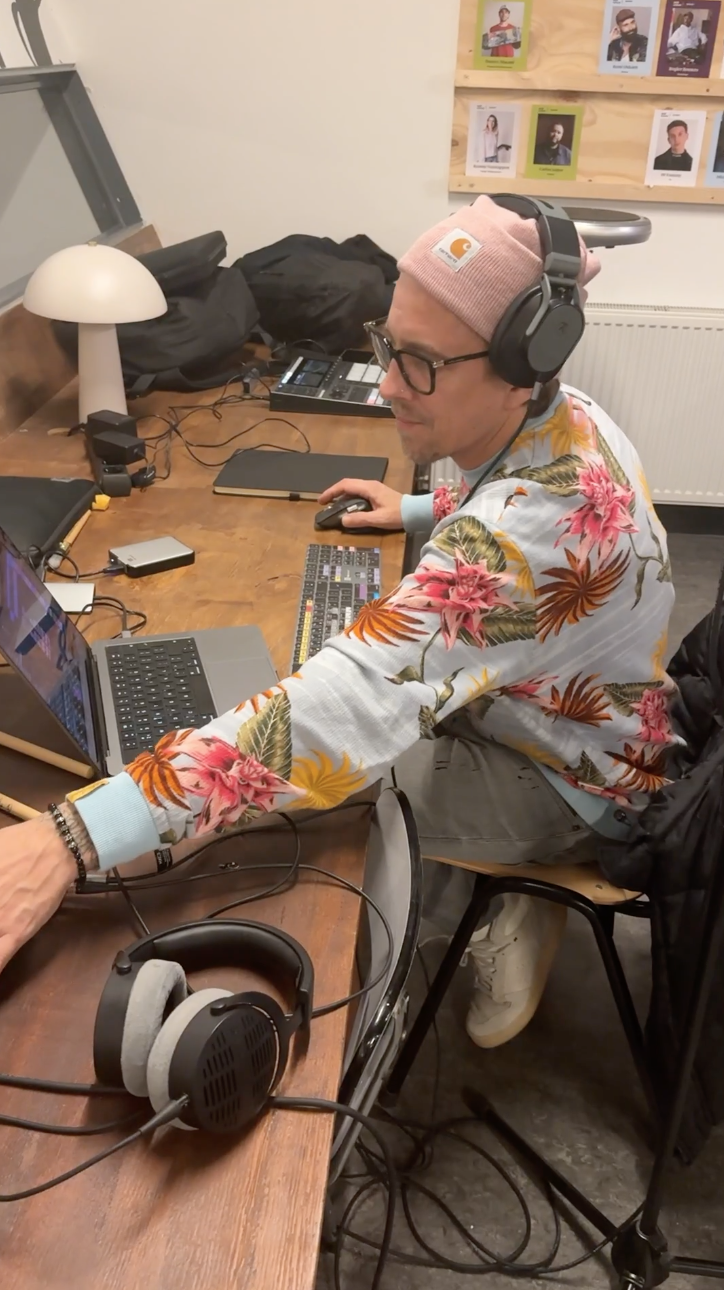 Man met een luide koptelefoon, laptop en geluidsapparatuur op een houten tafel, omgeven door technische apparatuur en een muur met foto's. Skill School leerlijn muziek maken in Leiden.