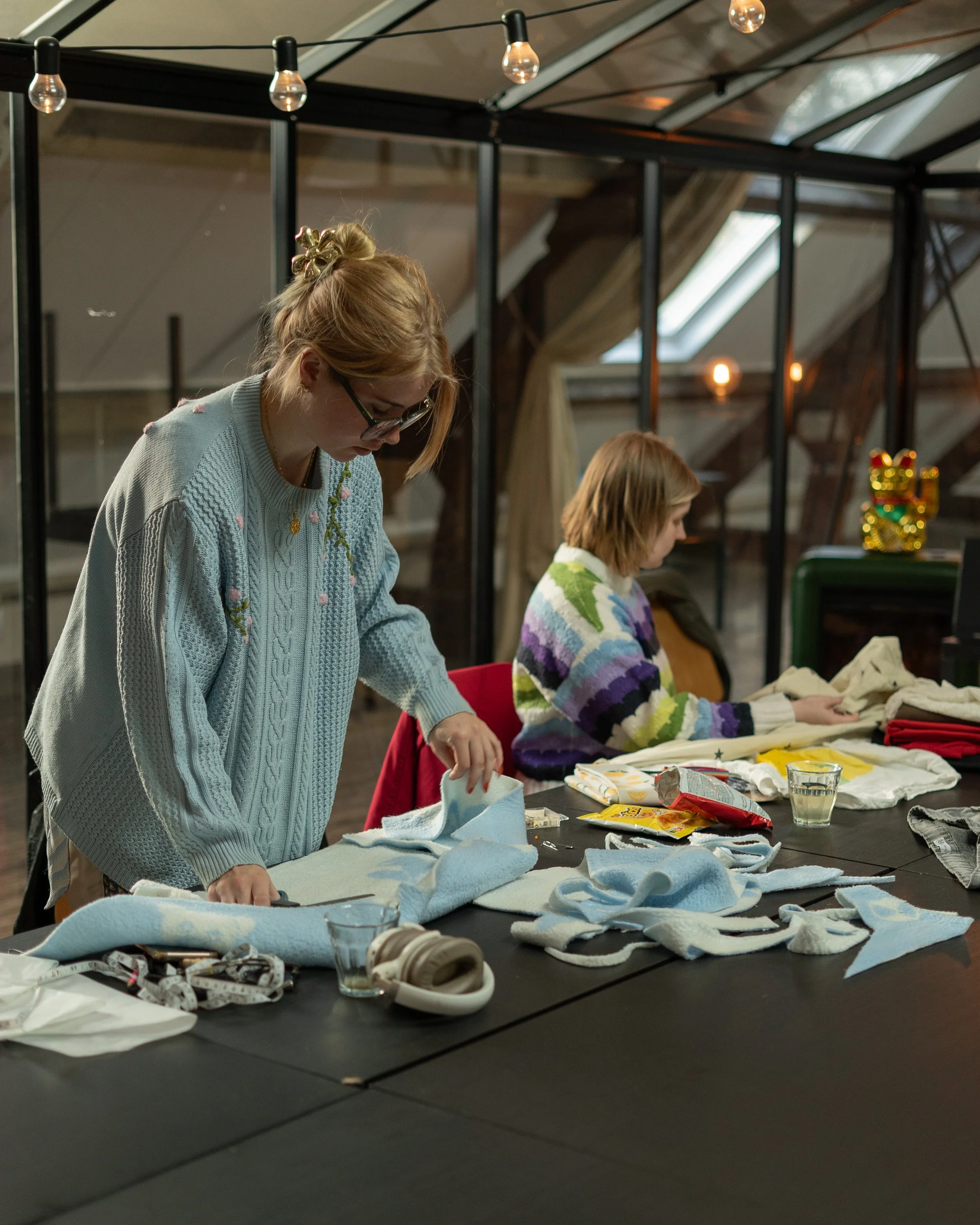 Twee vrouwen werken aan creatieve projecten aan een lange tafel met stoffen, scharen en meetlinten in een ruimte met veel natuurlijk licht. Kleding maken leerlijn bij Skill School