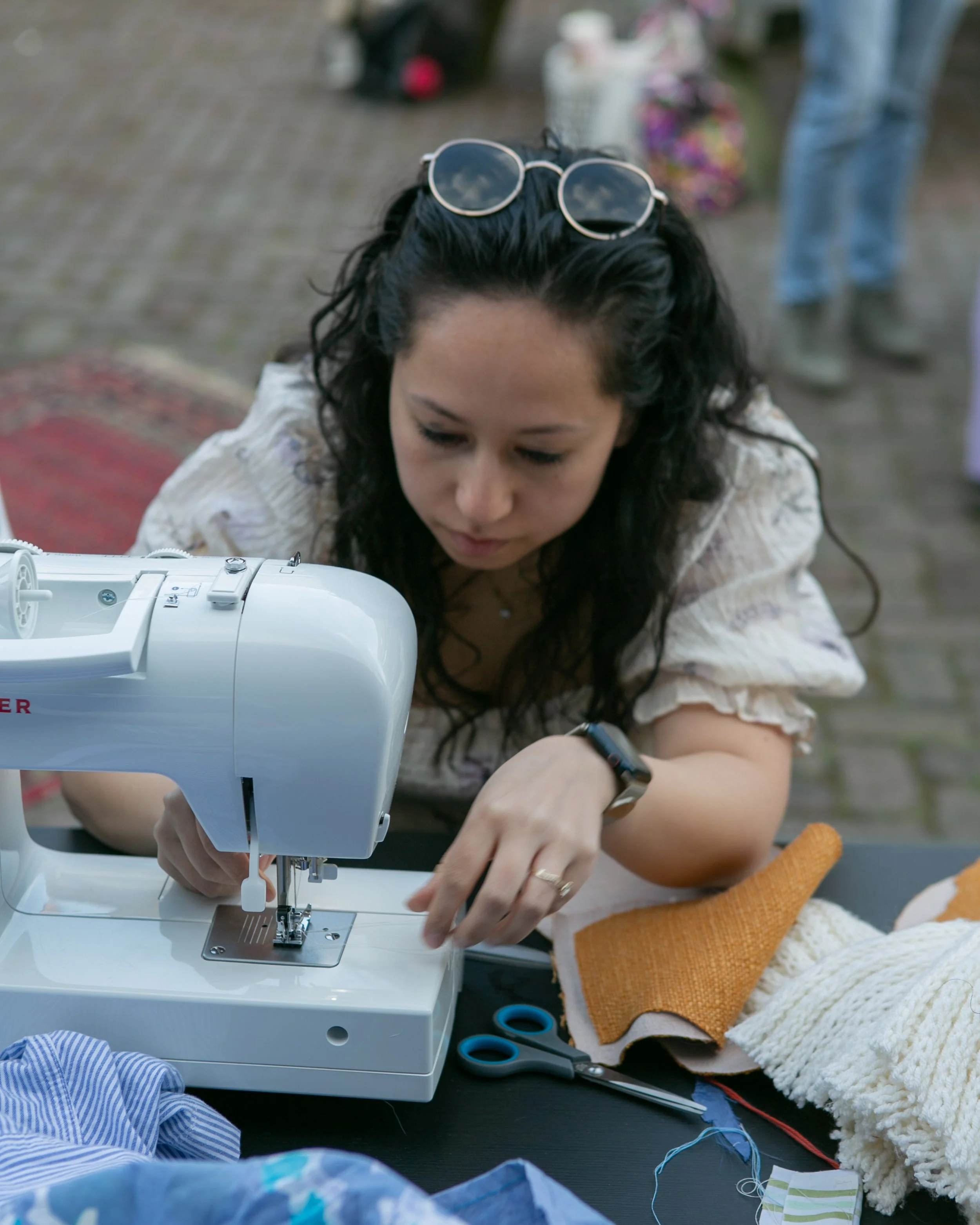 Vrouw met zwarte krullen en sunglasses op haar hoofd die een naaimachine bedient, omringd door stoffen en een paarse schaar buiten. Kleding maken leerlijn bij Skill School