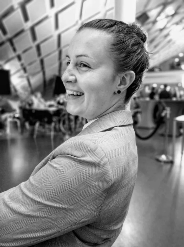 Een vrouw glimlacht en kijkt over haar schouder in een drukke, moderne binnenomgeving. Docent bij Skill School