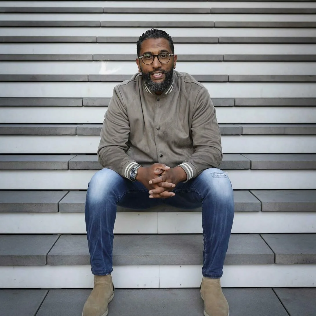 Man zit op trappen en kijkt naar de camera. Hiervan is de achtergrond een muur met horizontale lijnen. Skill School docent.