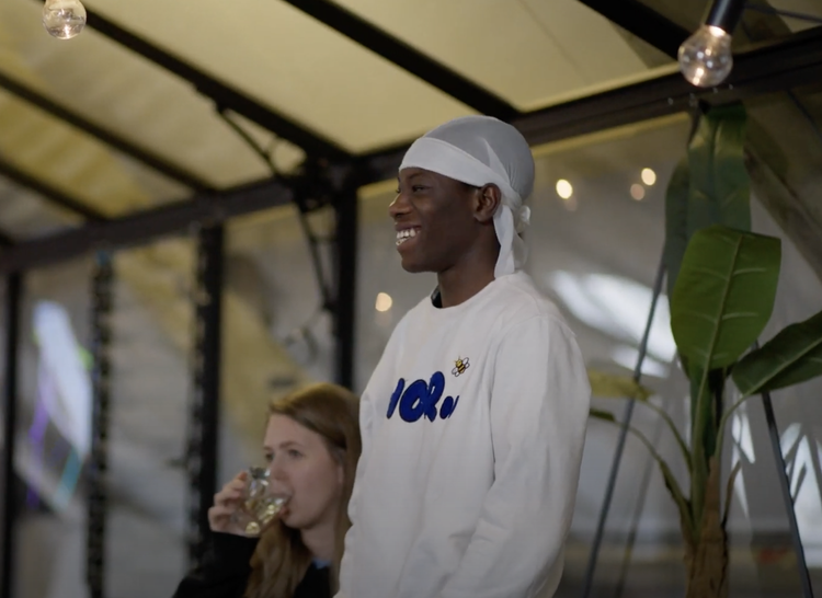 Een man lacht terwijl hij in een tent zit, met een vrouw die drinkt, er zijn decoraties en planten in de achtergrond. Skill School Ede.