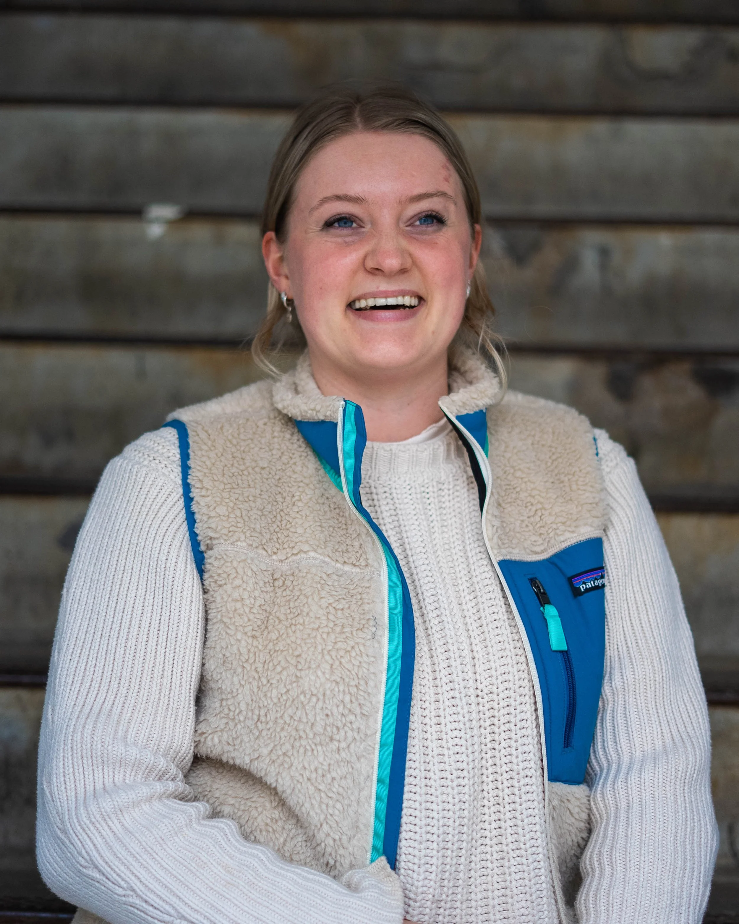 Een lachende vrouw met blond haar, gekleed in een witte gebreide trui en een blauwe outdoor jas, staat voor een houten achtergrond. Skill School docent.
