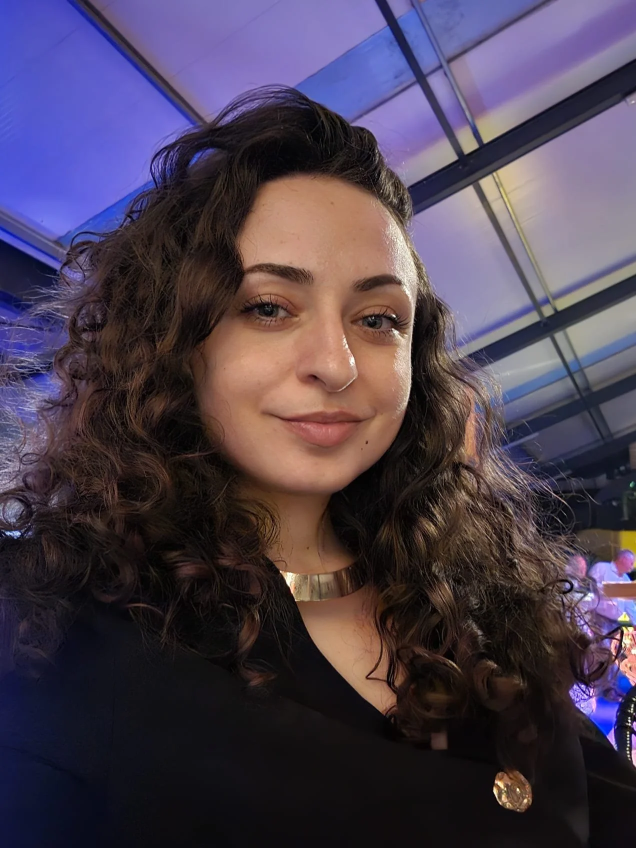 Een portretfoto van een jonge vrouw met lang, krullend haar in een lichte omgeving met blauwe en paarse verlichting. Docent bij Skill School