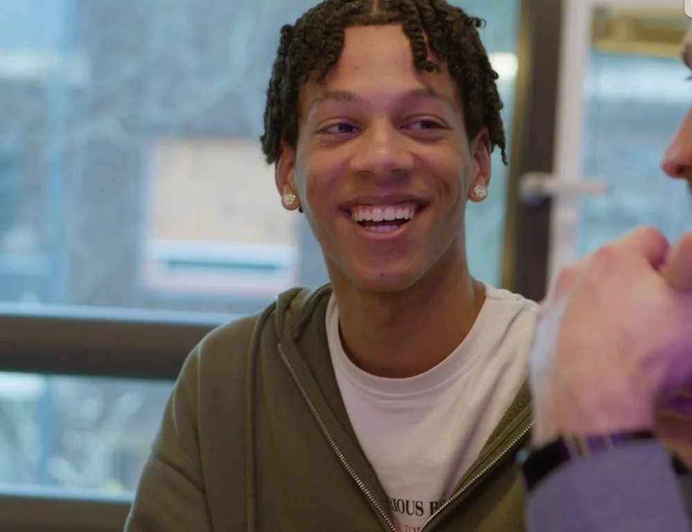 Een jonge man met kort krullend haar en oorringen die glimlacht tijdens een gesprek. Skill School jongere.