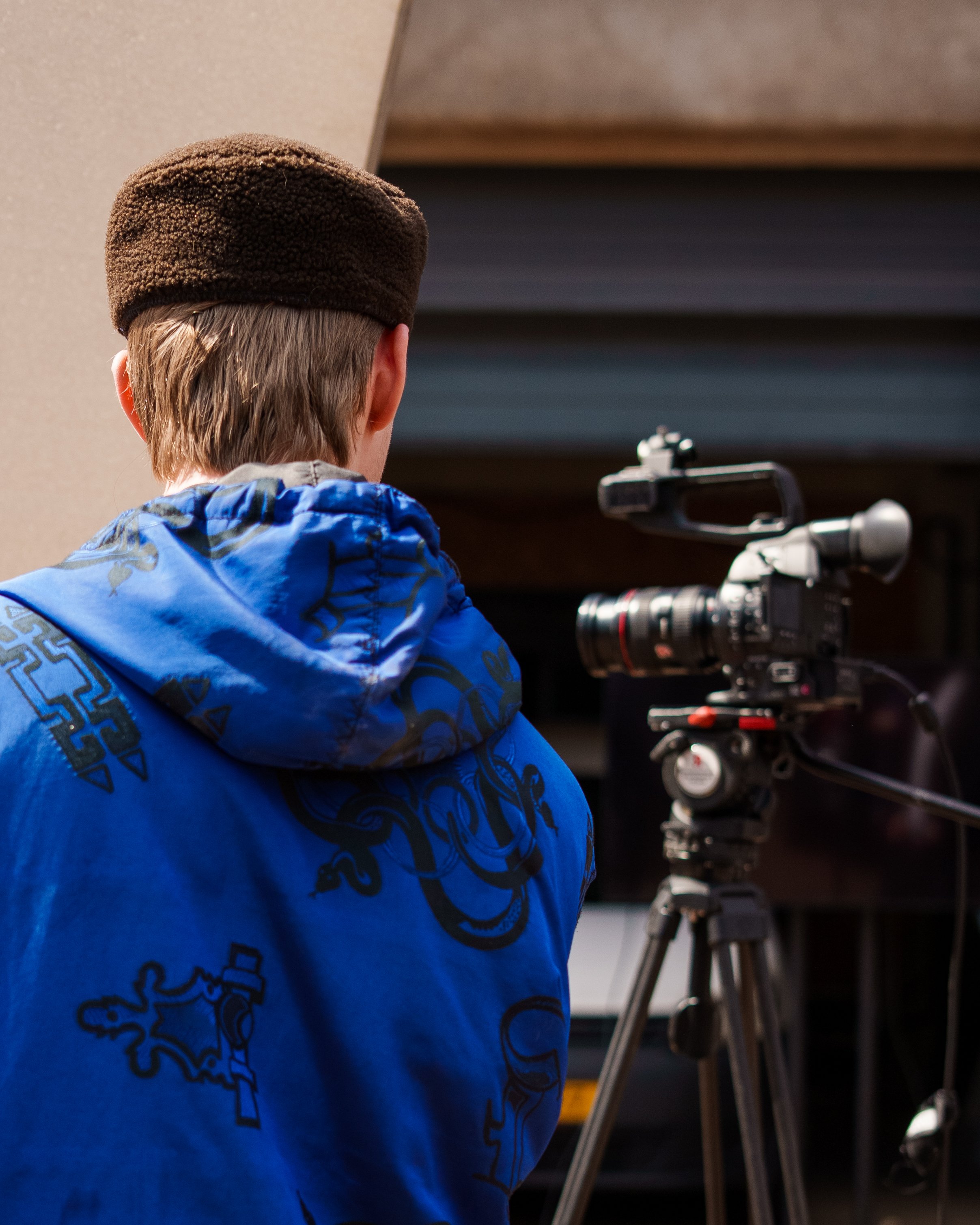 Jongen met pet en blauwe jas bedekt zijn gezicht achter een professionele camera op een statief. Filmen en editen bij Skill School