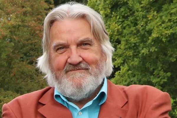 Een oudere man met grijs haar en een volle baard staat buiten, gekleed in een bruin jasje en een blauwe overhemd, met groene bomen op de achtergrond. Skill School docent.