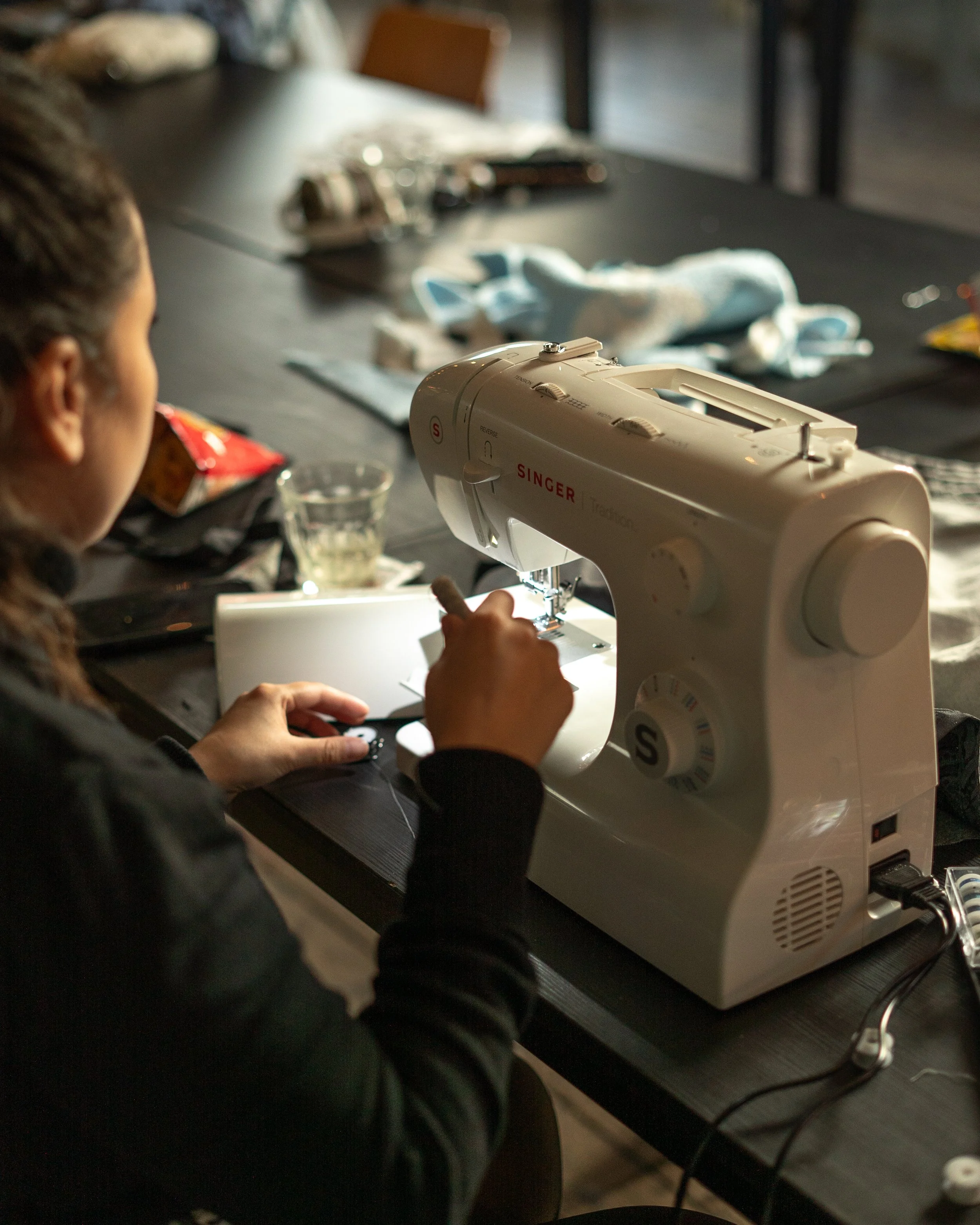 Een persoon die op een naaimachine kleding of stoffen naait op een tafel met verschillende items zoals snackverpakkingen, een glas en lapjes stof. Kleding maken leerlijn bij Skill School
