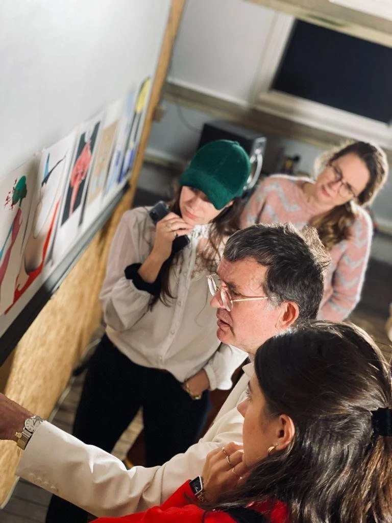 Vijf mensen kijken naar kunstwerken op een muur, een lijkt uitleg te geven, anderen luisteren aandachtig. Skill School docent.