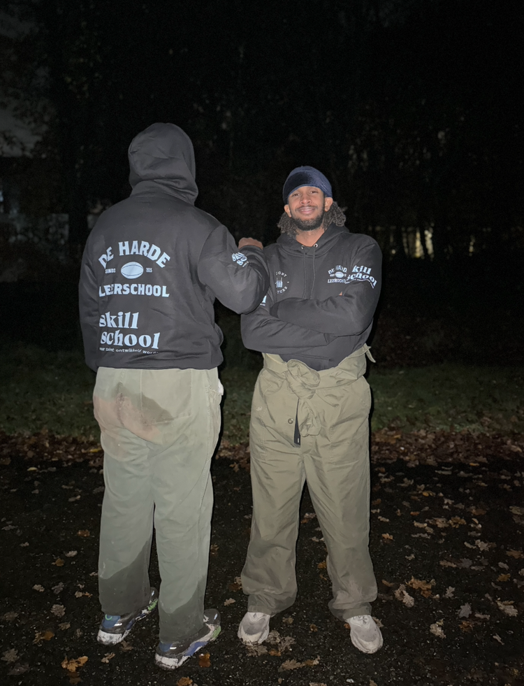 Twee mannen in sportkleding staan buiten 's nachts, een omhelst de ander waardoor alleen de achterkant zichtbaar is. Ze dragen zwarte jassen met logo's en slogans, en broeken. Skill School leerlijn Reset.