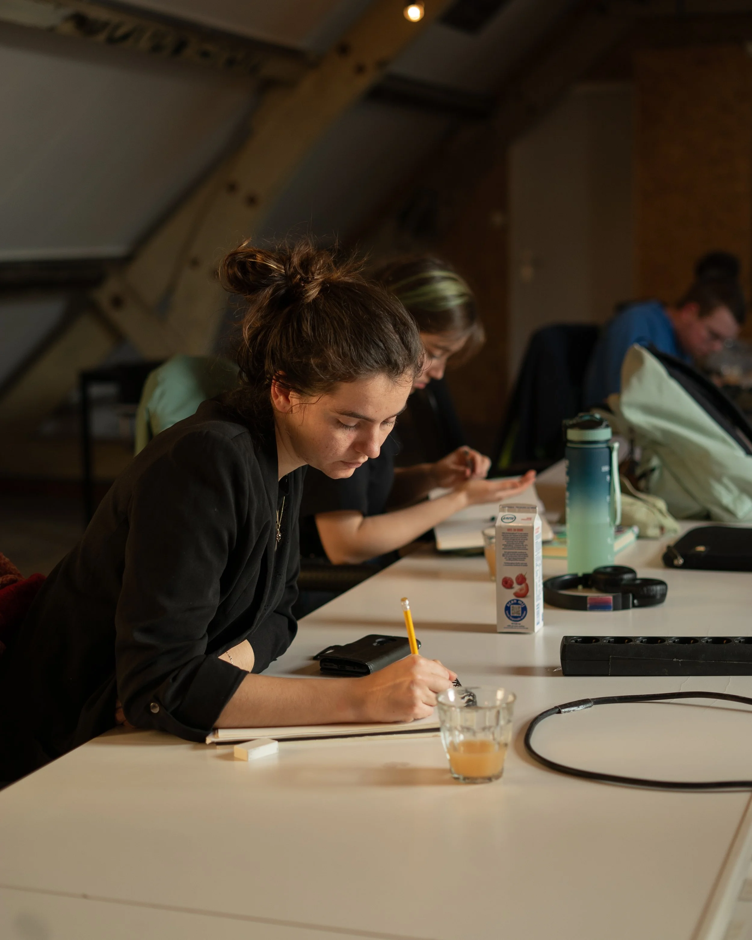 Vrouwen zitten aan een tafel, één schrijft in een notitieboekje terwijl een andere op haar smartphone kijkt, in een creatieve of werkruimte. Striptekenen en animeren bij Skill School