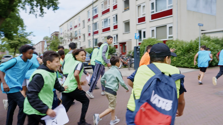 Groep jonge kinderen die buiten in een woonwijk rennen en spelen, sommigen dragen kleurige sport- of speelvesten. Skill School The hunt.
