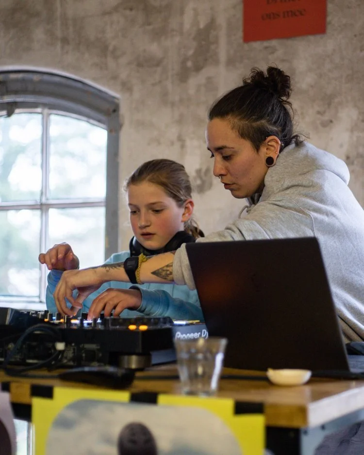 Een vrouw en een meisje werken samen aan een dj-setup, met een laptop op tafel, in een kamer met een bakstenen muur en een raam. Skill School docent muziek maken.