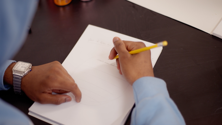 Persoon schrijft op een vel papier met een gele potlood, op een donkere tafel. Skill School leerlijn muziek maken. Hij maakt zijn eigen lyrics.