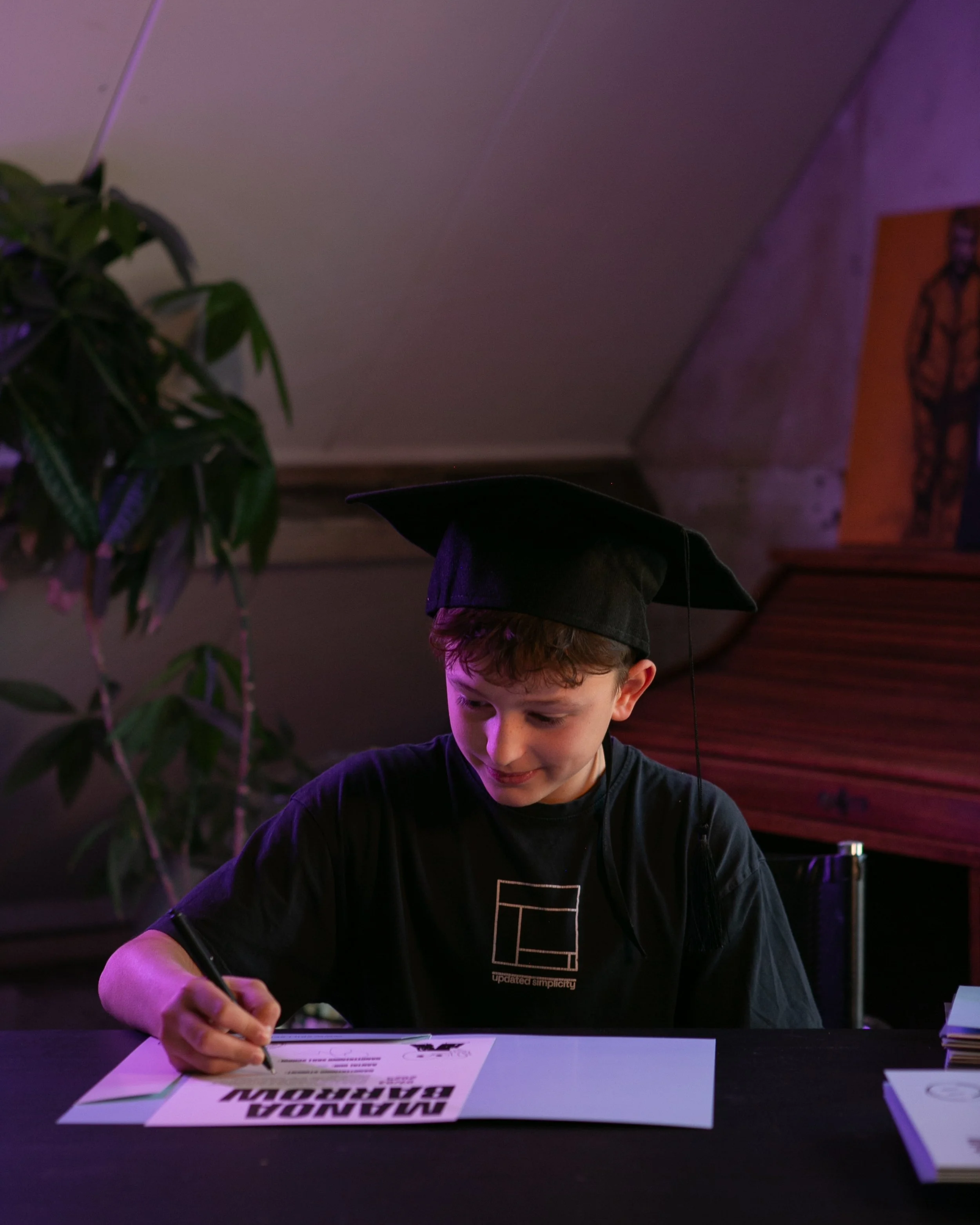 Jongen met een zwart afstudeerhoed die op een tafel ondertekent. Skill School afstudeer festival.