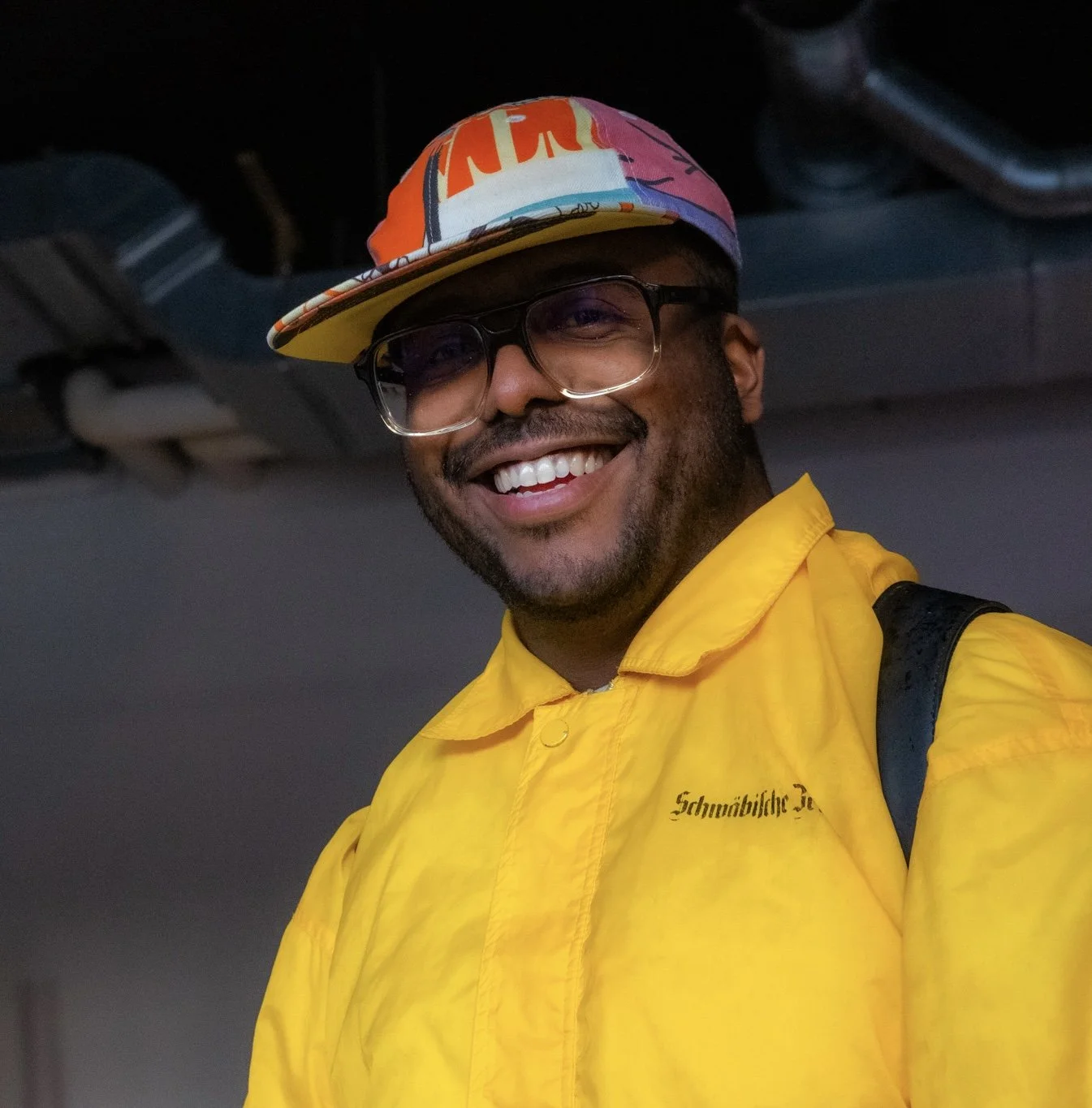 Man met donkere huid, glimlachend, draagt een gele jas, een gekleurde pet, en zwart montuur bril, binnen. Docent bij Skill School Ede.