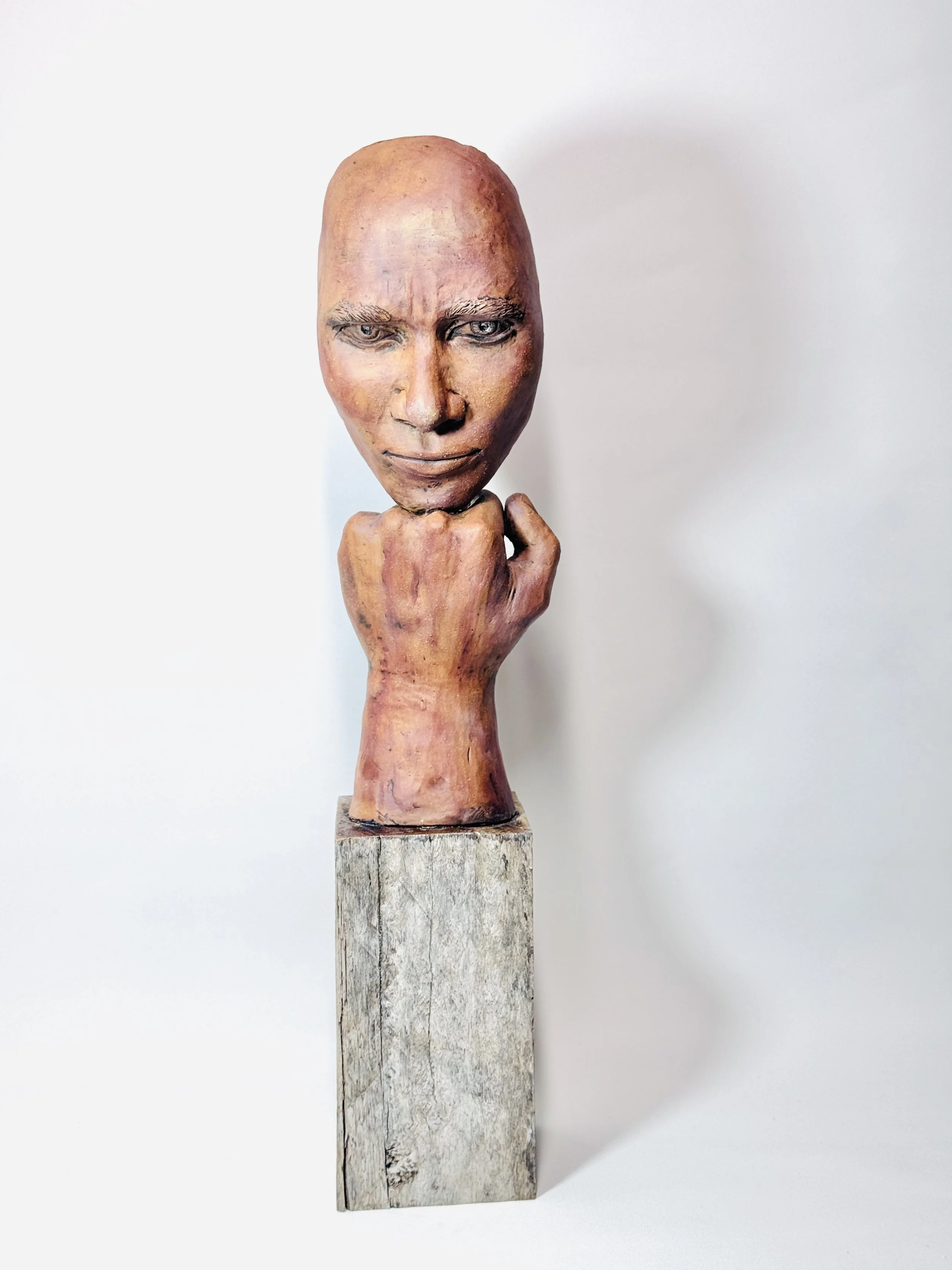 A wooden sculpture of a human head with detailed facial features and a contemplative expression, mounted on a tall, weathered wooden pedestal.