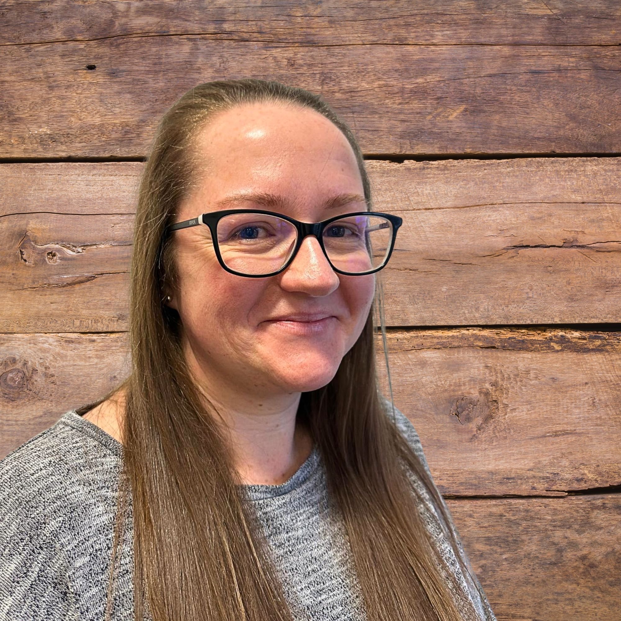 A woman with long brown hair and glasses, smiling, in front of a wooden background.