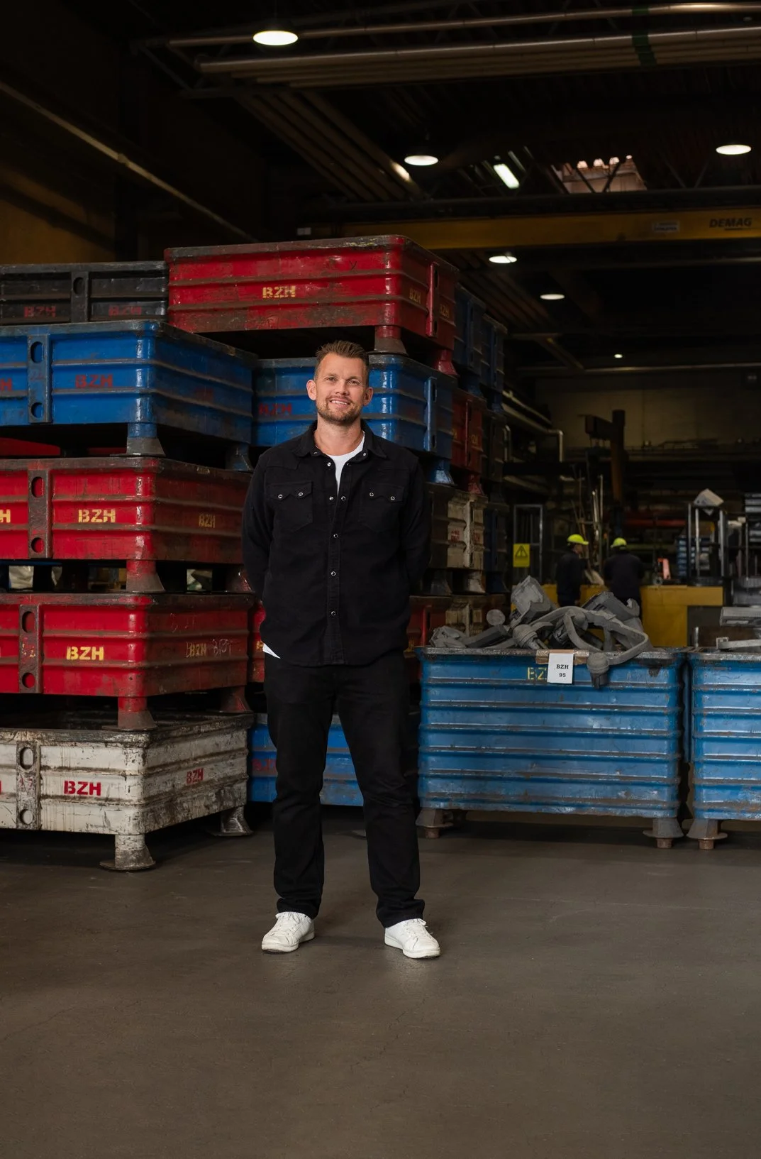 A man dressed in black standing in front of large industrial storage bins in a warehouse with safety workers in the background.