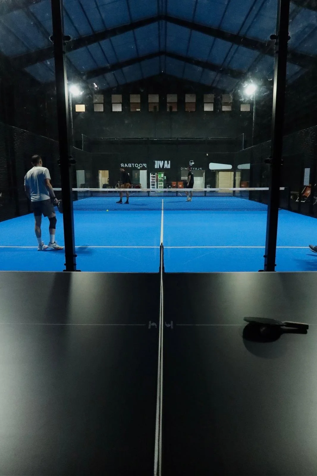 Vue à travers une vitre d'une salle de tennis de table avec deux joueurs en train de jouer et deux autres dans le fond.