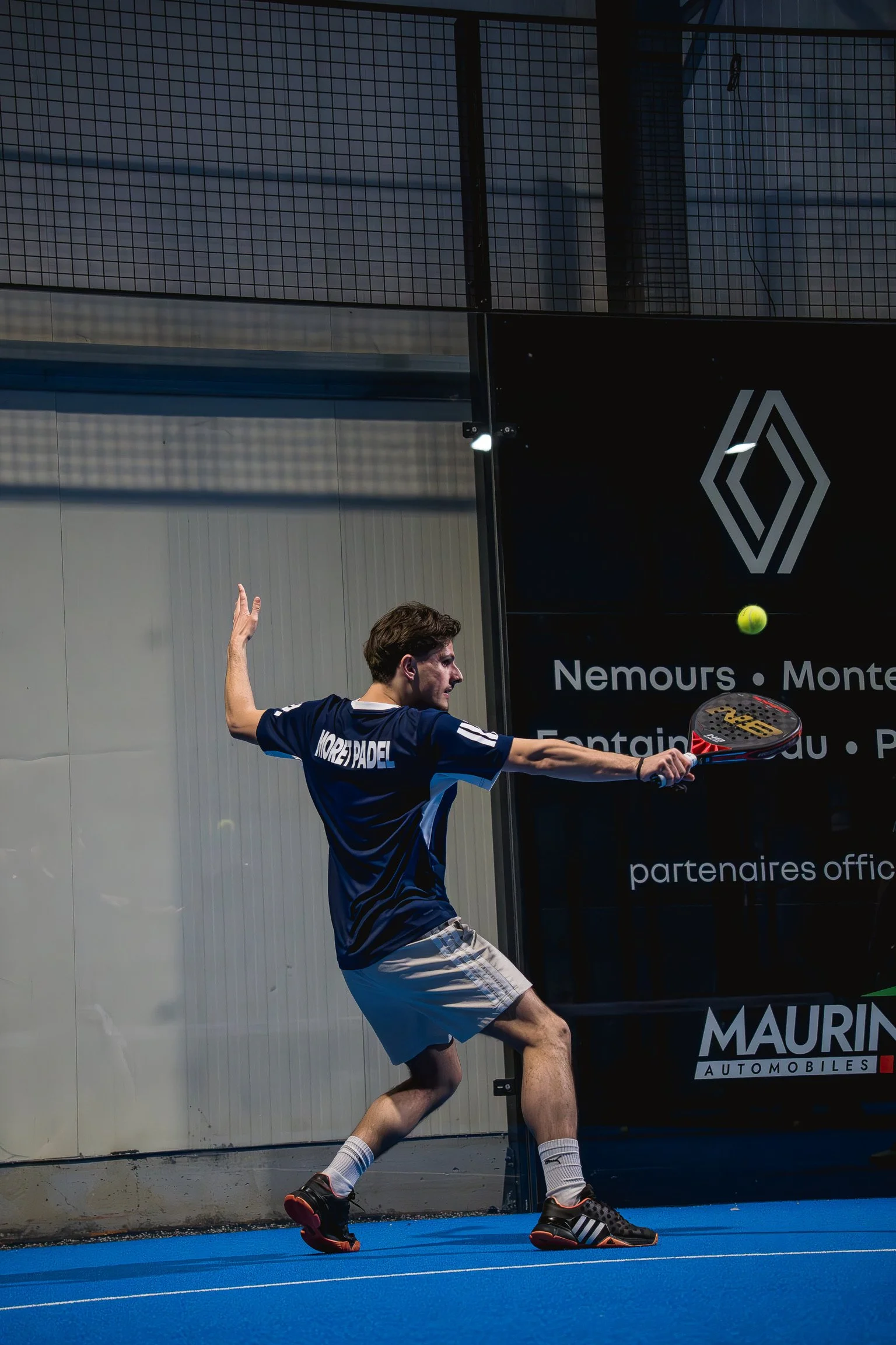Un homme joue au padel en frappant une balle avec une raquette. Il porte un t-shirt bleu, un short gris, et des chaussures de sport. La scène se déroule à l'intérieur, sur un terrain de padel avec un mur en arrière-plan et un panneau publicitaire.