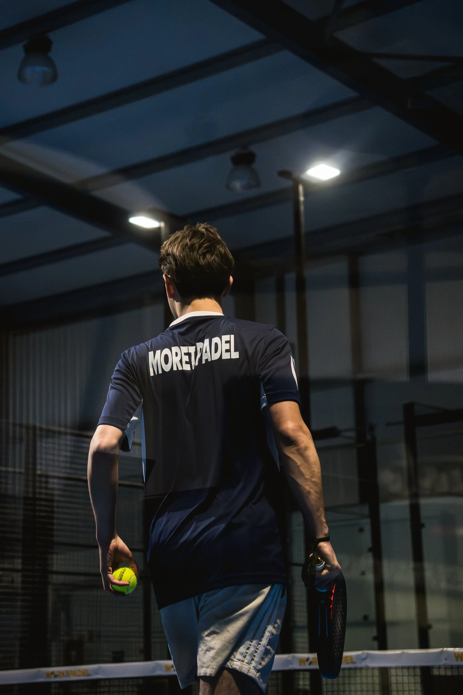 Un jeune homme de dos, tenant une balle de tennis dans sa main droite et une raquette dans sa main gauche, sur un court de tennis intérieur sous un éclairage artificiel, portant un maillot noir avec le nom 'Moret-ADEL' à l'arrière.