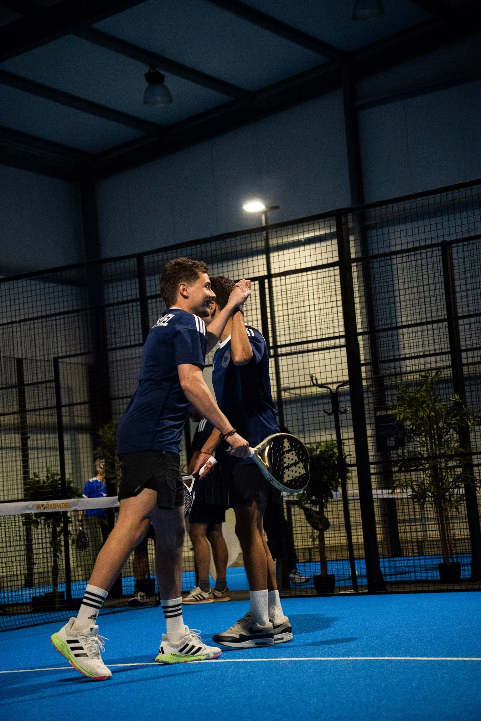 Deux jeunes hommes en tenue de tennis de table se félicitent dans une salle de sport en soirée, avec un filet de ping-pong et d'autres joueurs en arrière-plan.
