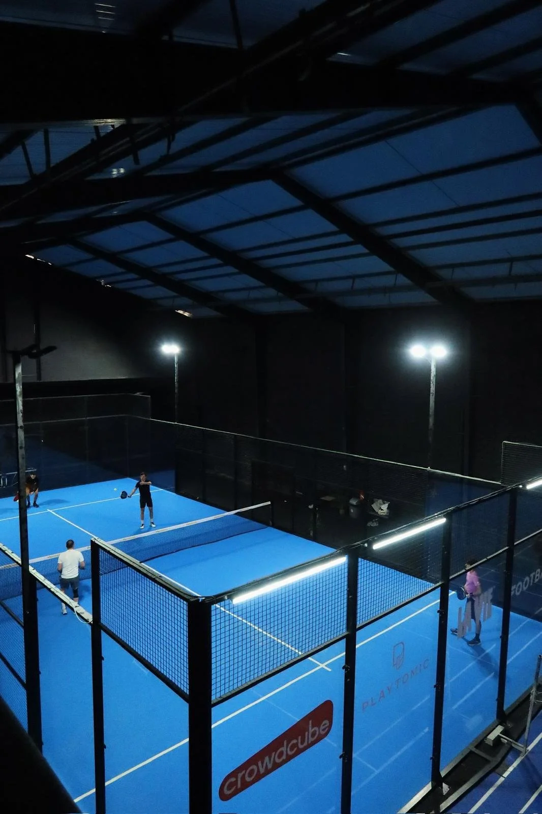 Une salle de sport intérieure avec un court de paddle-tennis éclairé, plusieurs personnes y jouent.