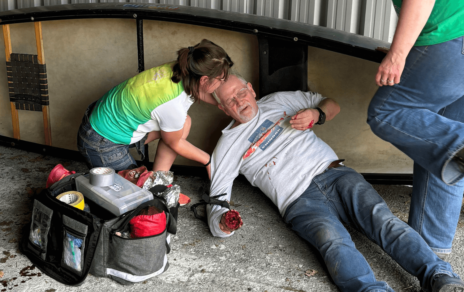 Person simulating a first aid scenario on a 16 hour outdoor first aid course lying on the ground with an arm wound and blood, receiving assistance from two other people, with first aid supplies nearby.