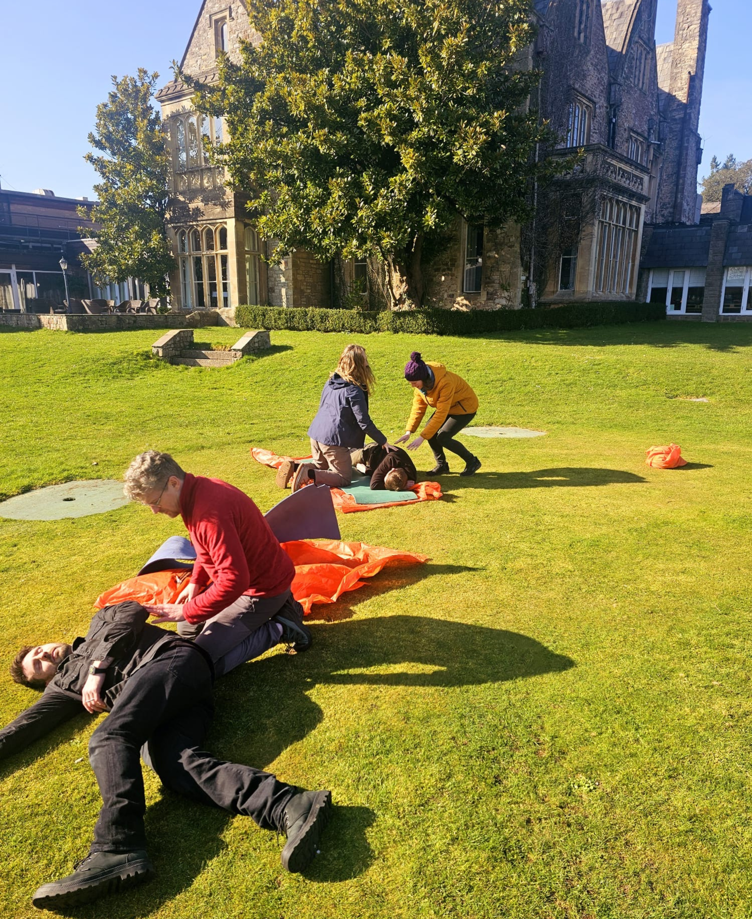 Group of people practicing first aid or CPR on a grassy lawn outside a historic building with large trees.