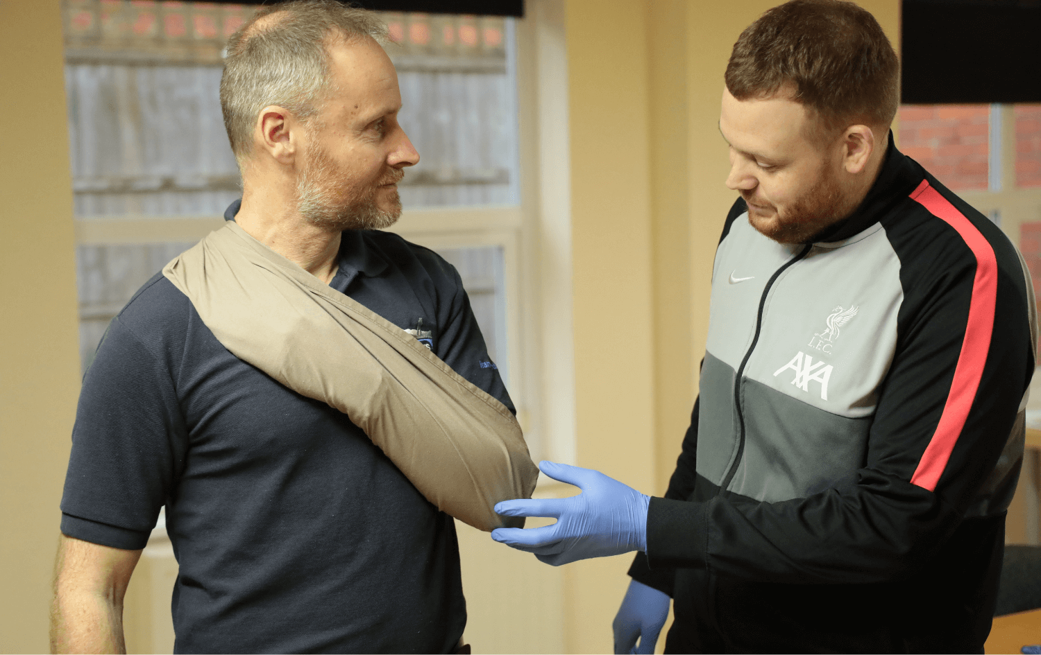 A man with a sling on his arm is being attended to by another man wearing a jacket with medical gloves, in an indoor setting on a first aid at work course.